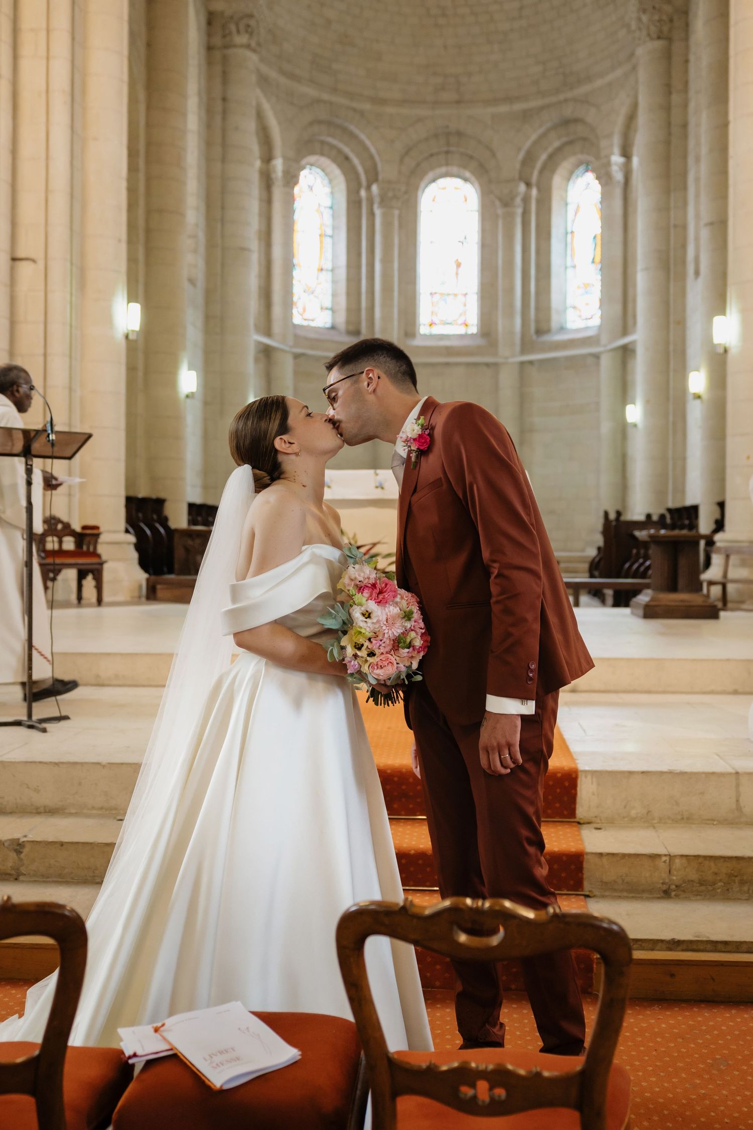 Bride and groom share a kiss at the altar in a church, bride in white gown holding pink bouquet, groom in burgundy suit.