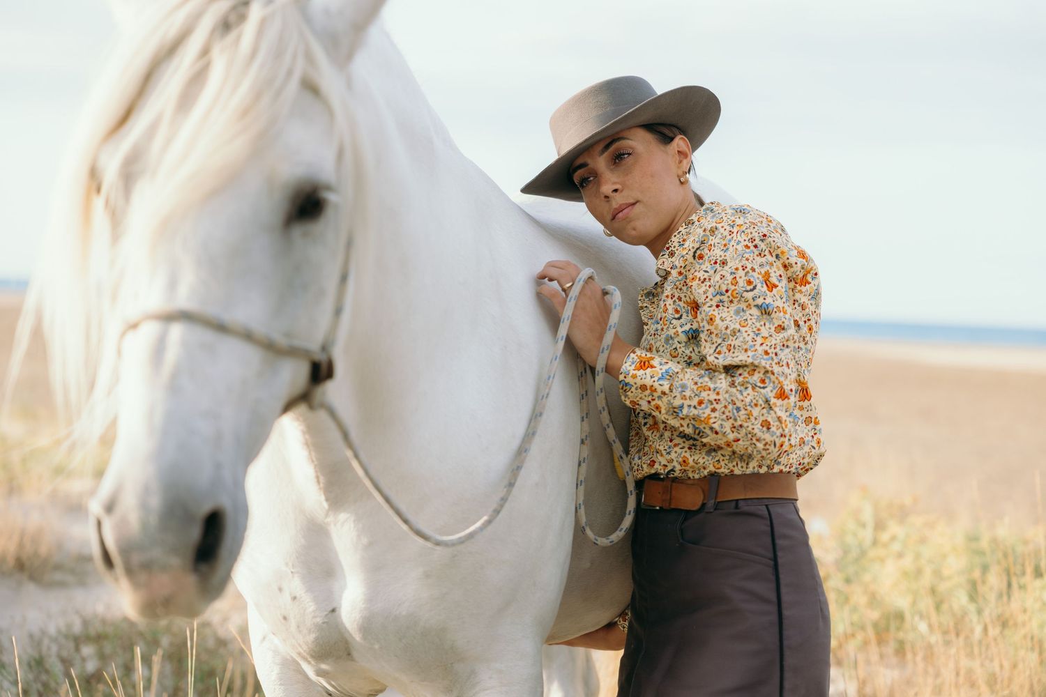 A white horse and rider in western attire stand together in a golden field during sunset.