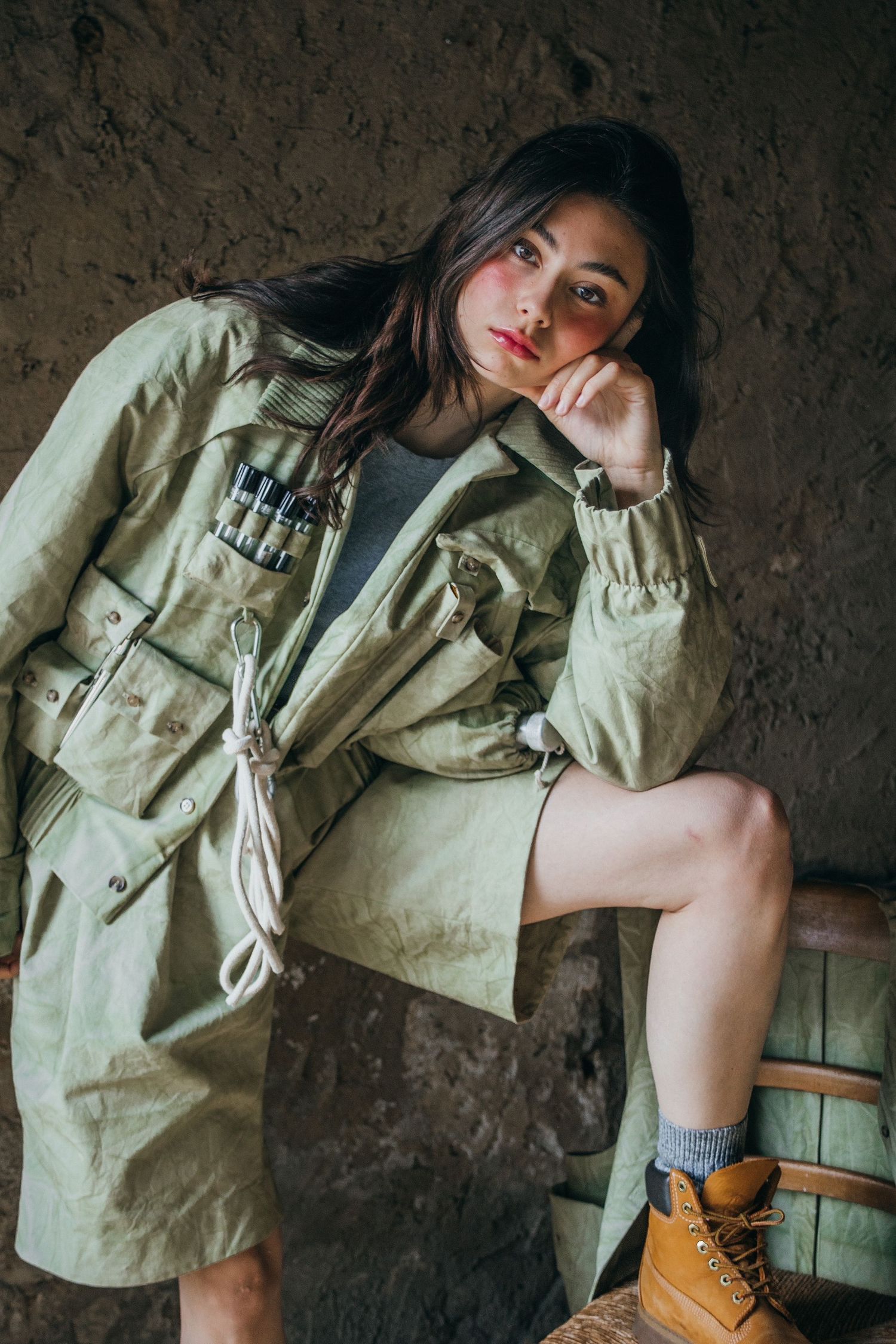 Stylish pose in an oversized military jacket and boots against a rustic wall.