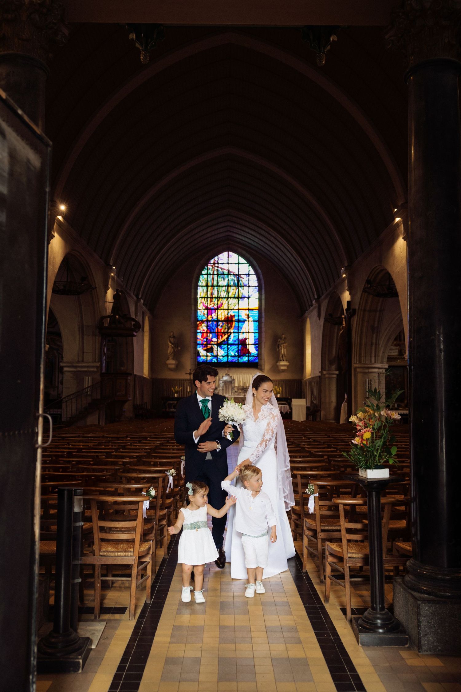 Wedding couple walks down church aisle with flower girls, stained glass window backdrop.