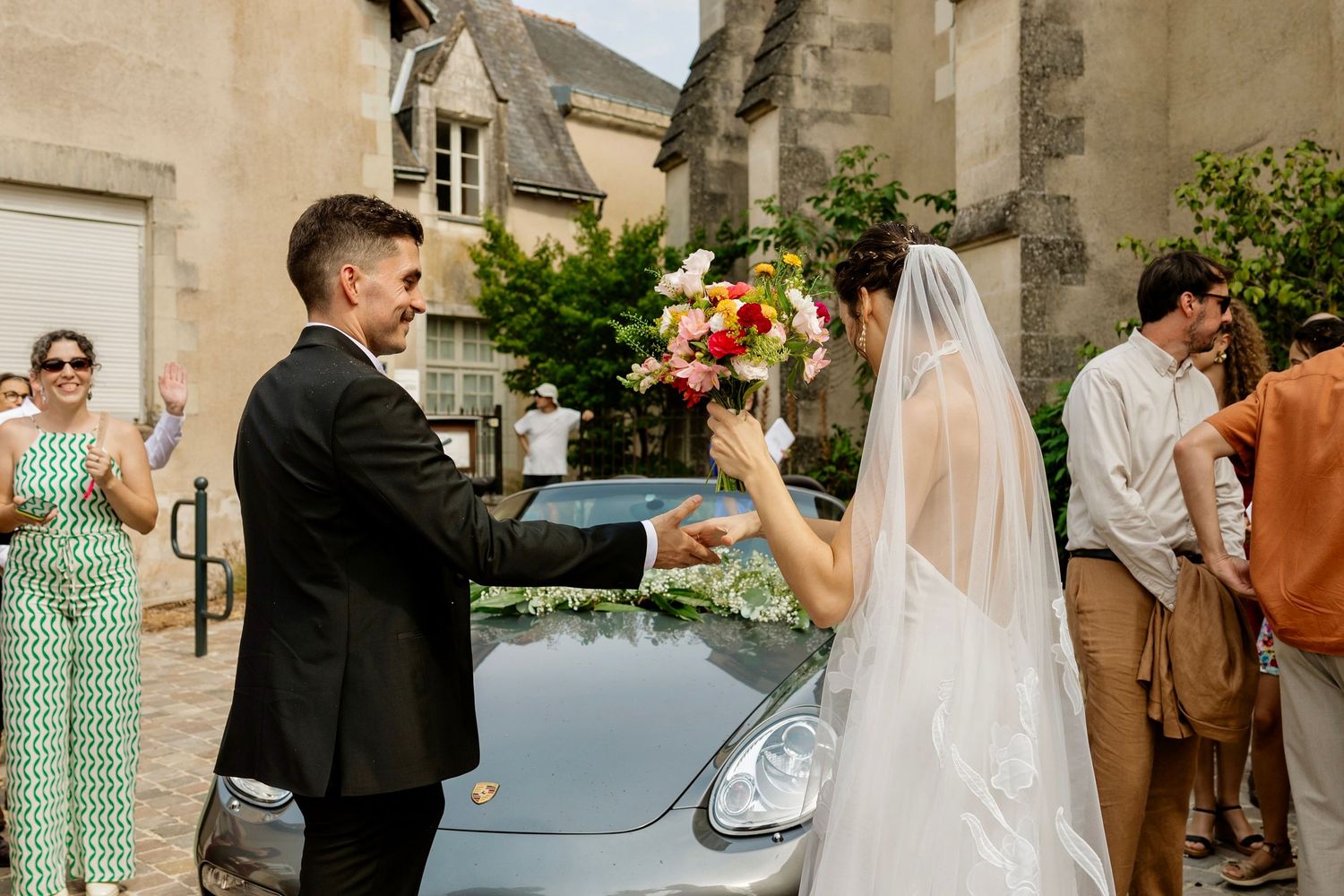 Bride and groom exchange bouquet beside a Porsche at an outdoor wedding ceremony in France.