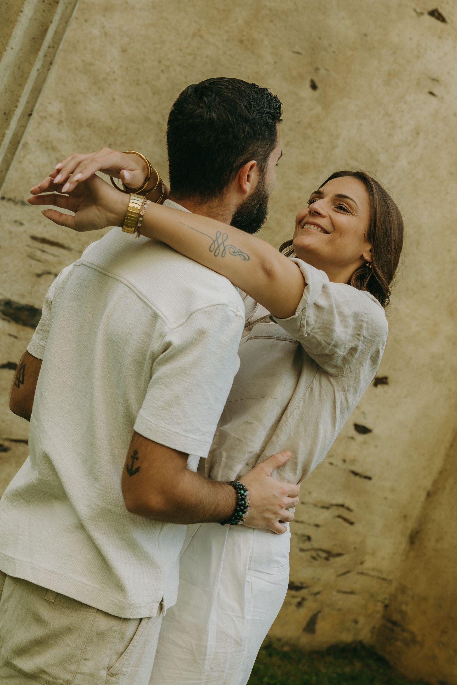 A couple in white embracing and smiling against a textured wall background.