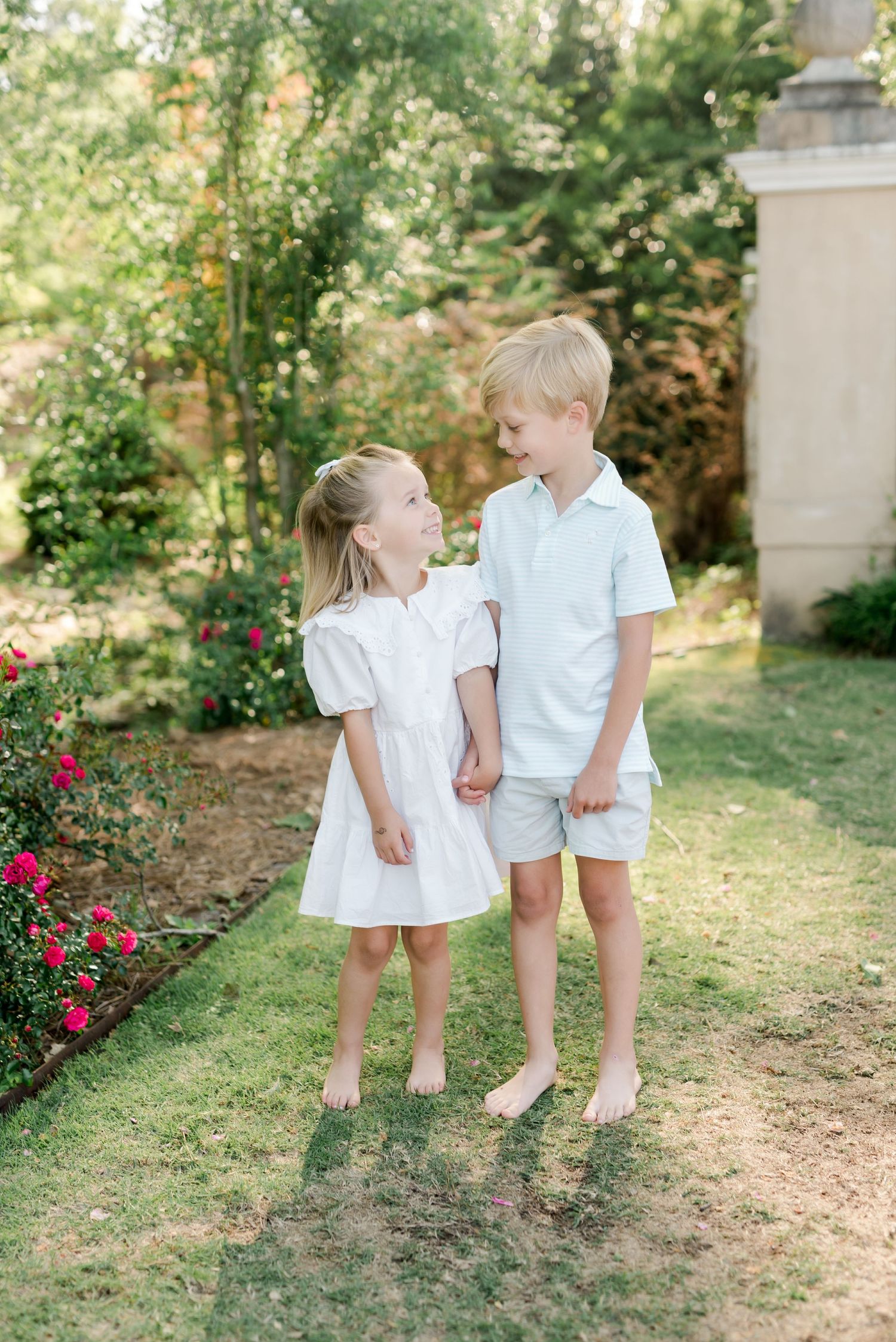 Two children in white summer outfits standing barefoot on grass near blooming rose bushes.