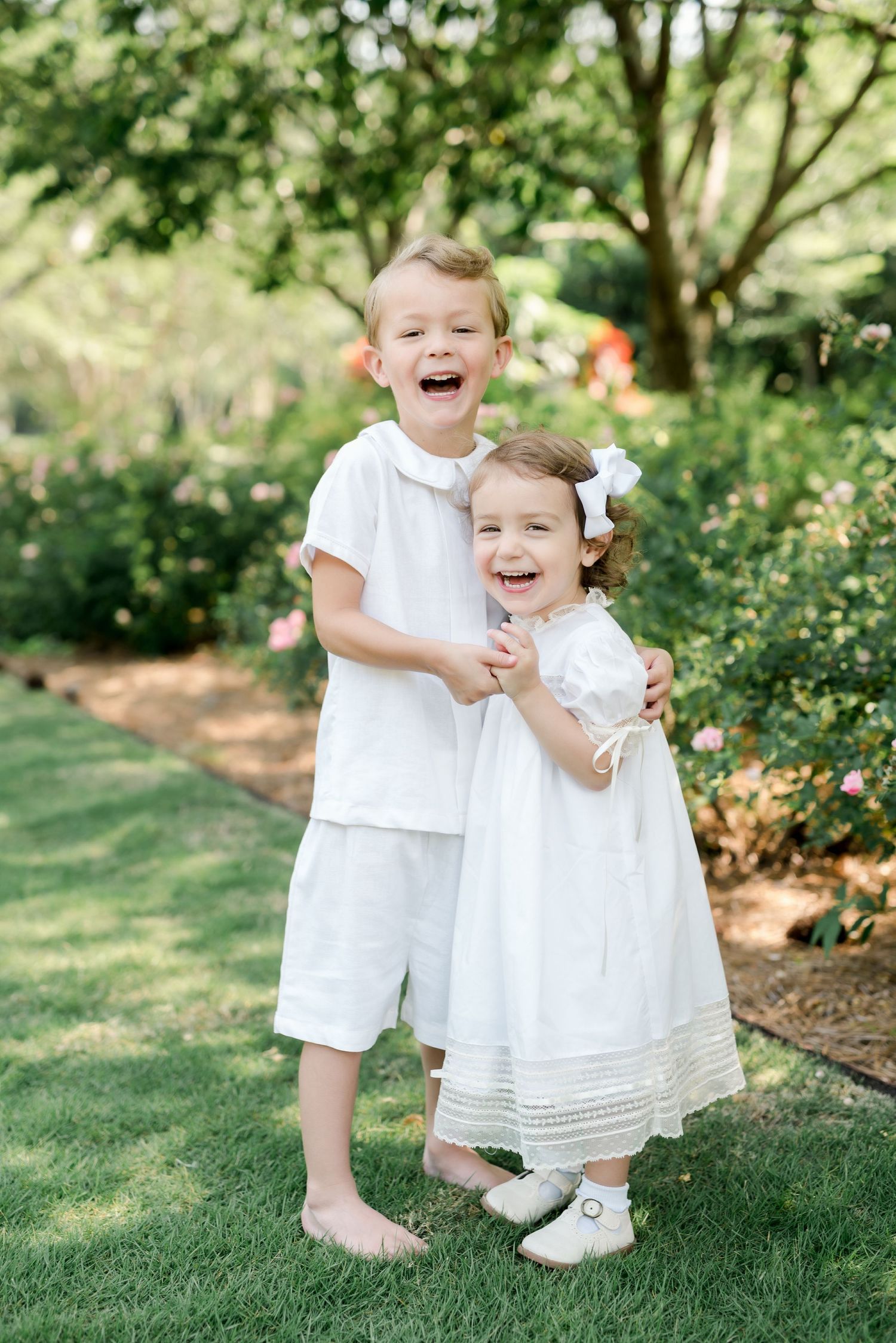 Two children in white outfits share a joyful embrace while standing in a lush garden setting.