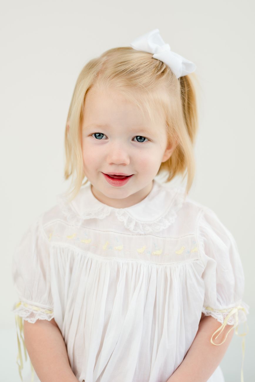 Child in white dress with bow and blonde hair smiling at camera against light background.