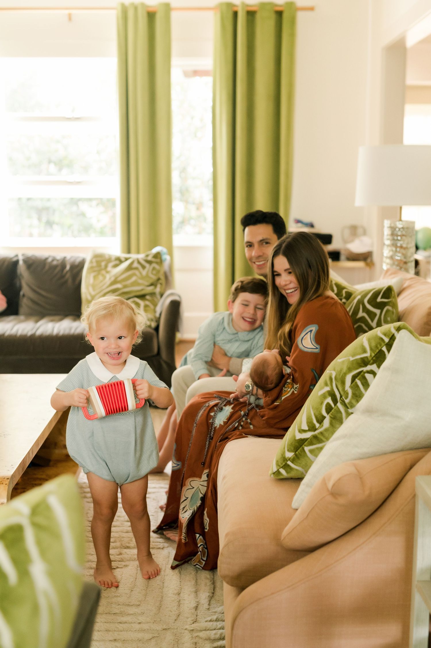 A happy family relaxes in a bright living room as a toddler in a blue romper plays with a red toy.