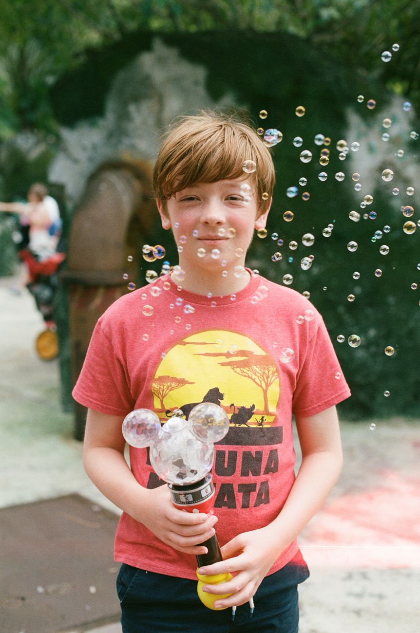 A smiling youth in a red t-shirt plays with soap bubbles in a theme park setting.