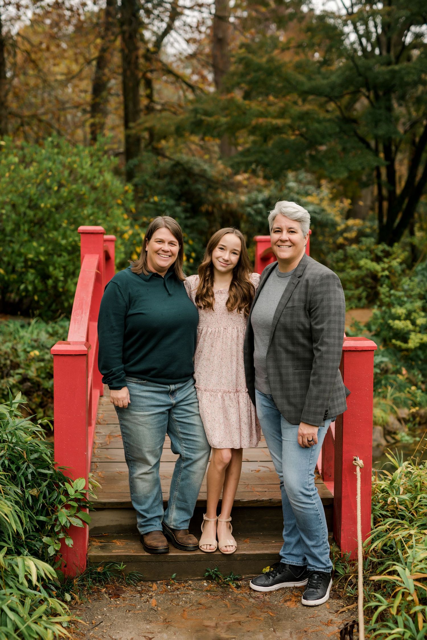 Three smiling people pose on a red wooden bridge surrounded by lush greenery in an autumn garden setting.