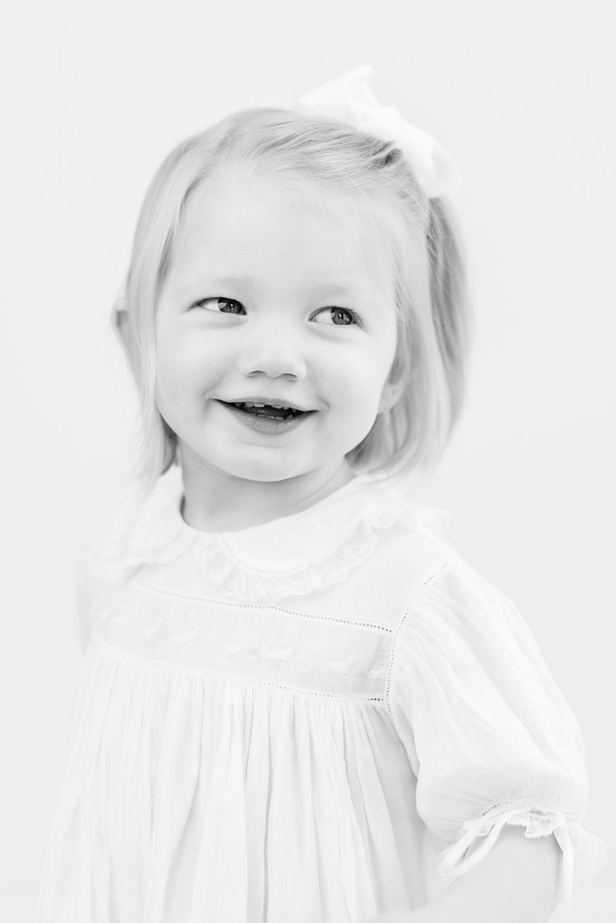 Black and white portrait of child in white dress with bow smiling brightly.