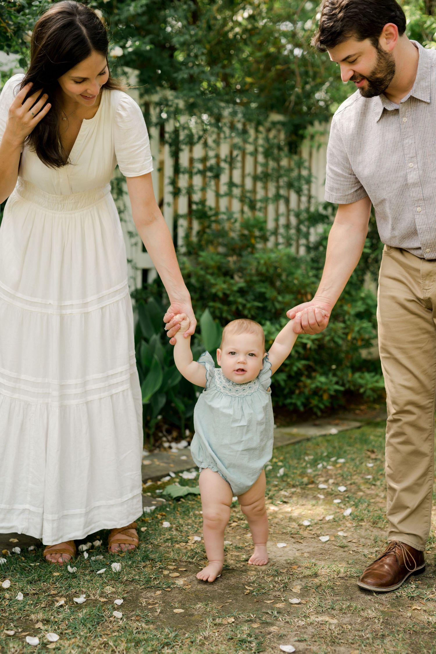 Parents helping baby take first steps in garden setting while wearing summer attire.