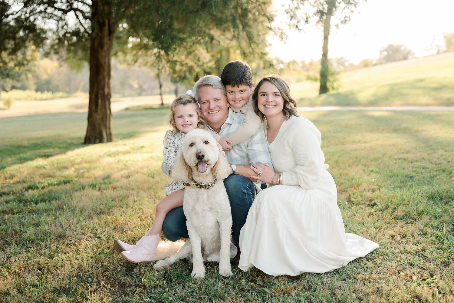 A fluffy white dog sits with a family on lush green grass during a golden hour photo session.