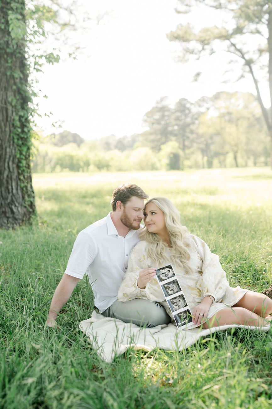 Couple sits on blanket in sunlit meadow holding ultrasound photos.
