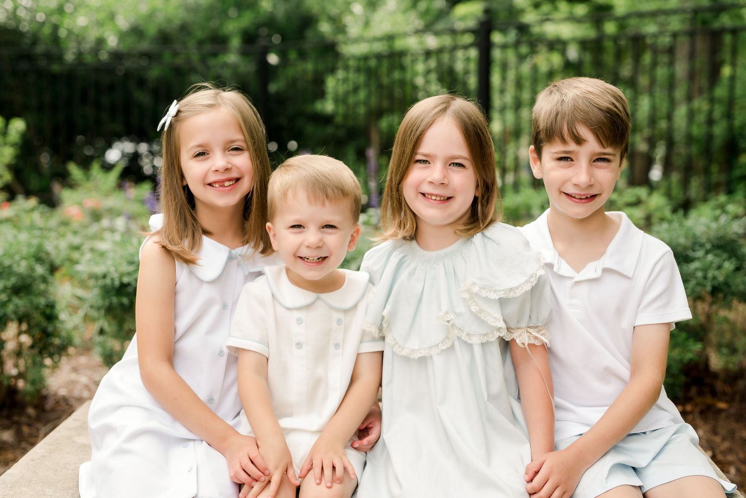Children in white outfits sit together for formal portrait in garden setting.