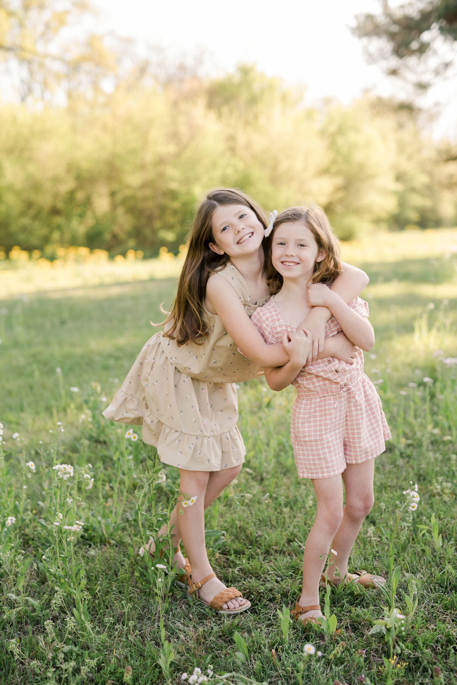 Two children in pastel dresses hug and laugh together in a sun-drenched meadow.