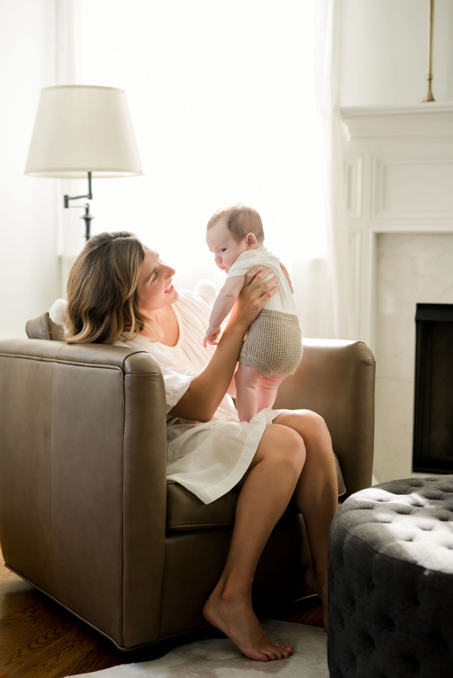 Mother lifts baby while sitting on brown leather chair in bright living room with white lamp.