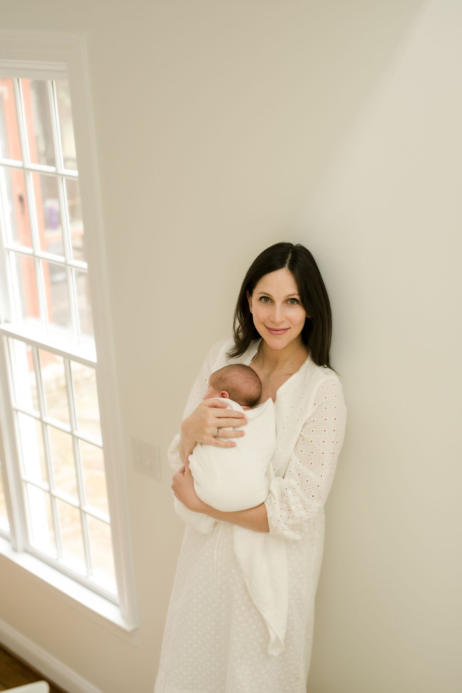 A mother in a white lace dress holds a newborn baby while standing next to a bright window in a minimalist room.
