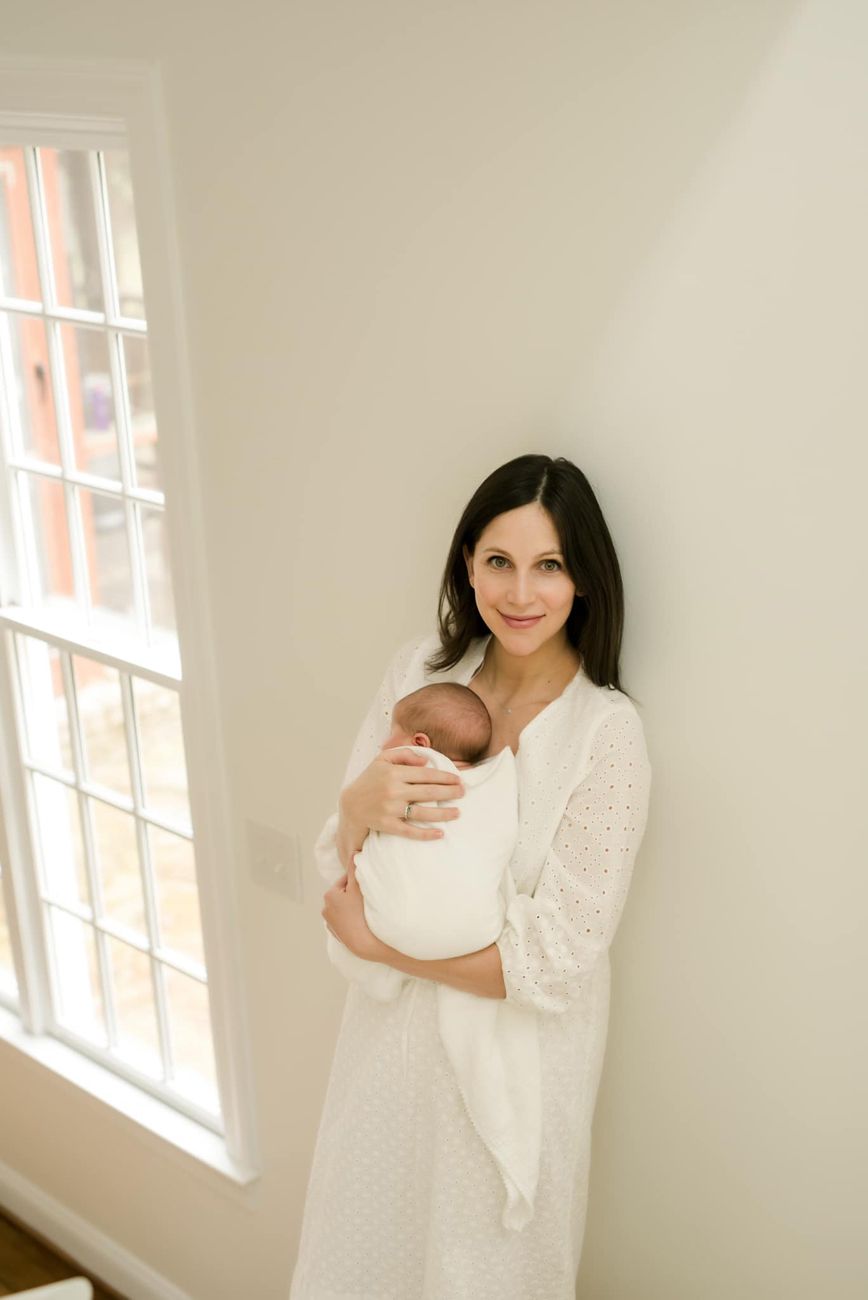 Maternity photoshoot in a bright white room with soft natural window light and flowing white lace dress.