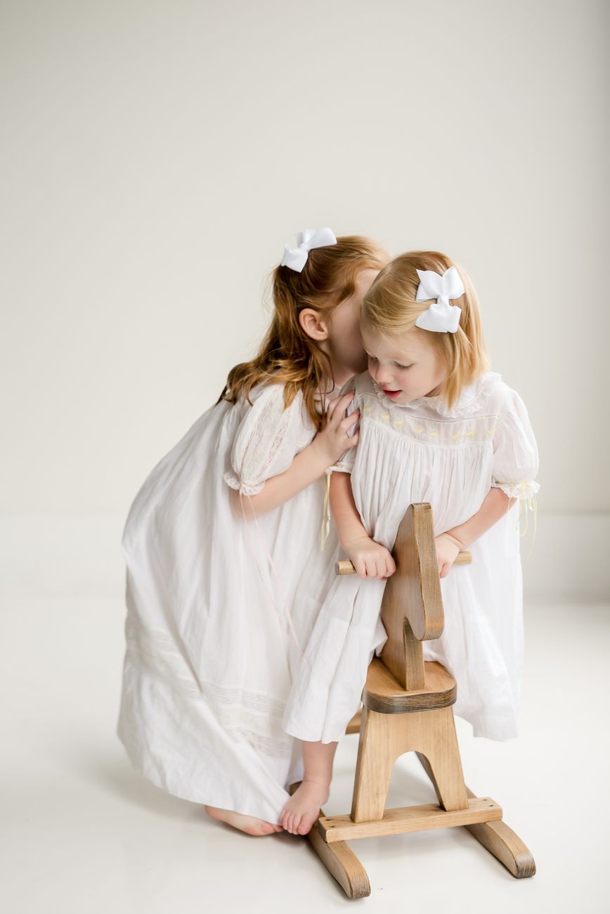 Two children in white dresses sharing a secret while playing with wooden rocking toy.