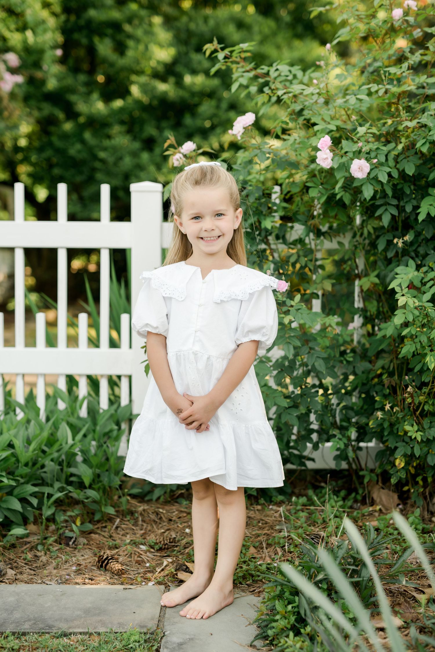 Young girl in white dress poses barefoot by white picket fence and rose garden.