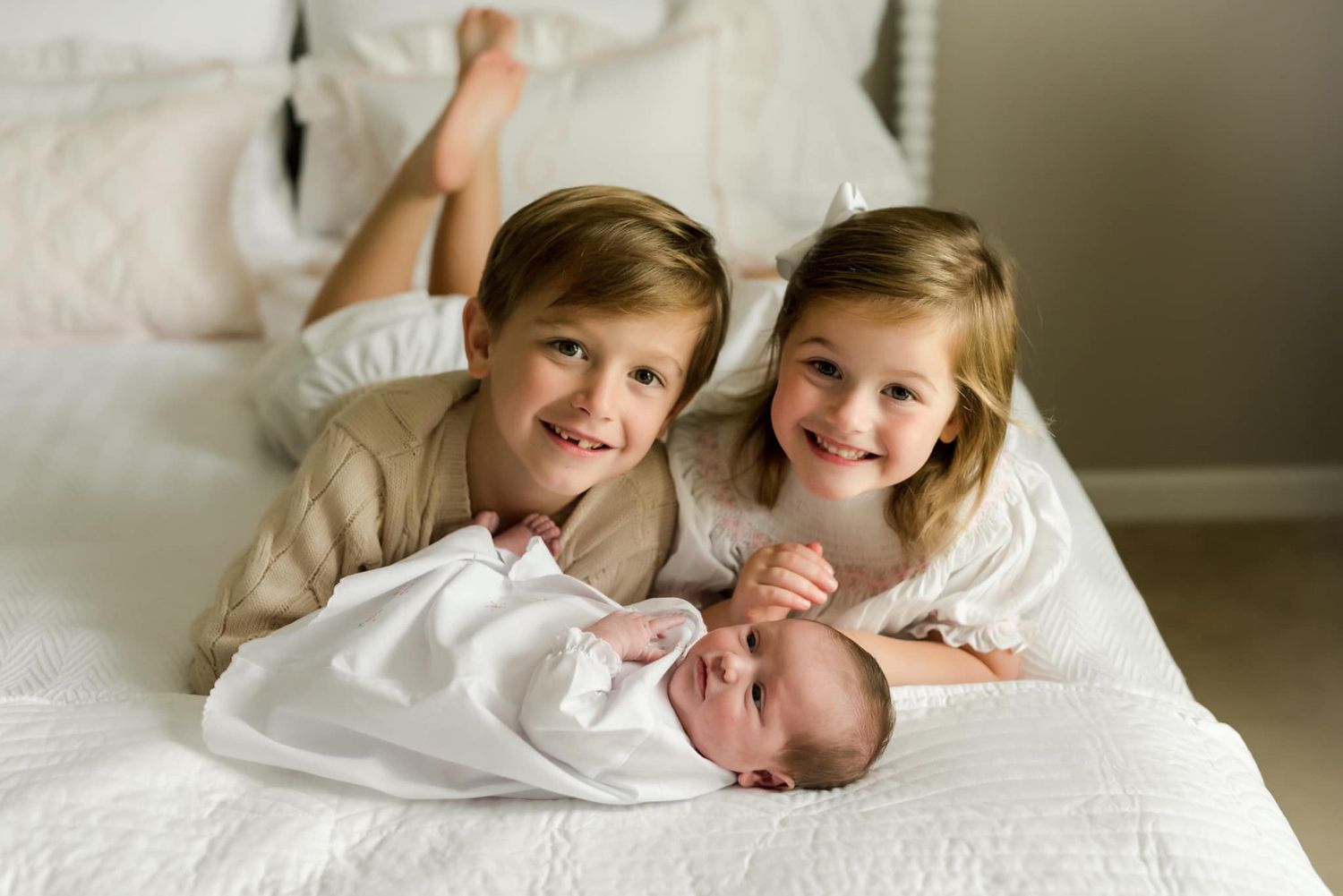 Two siblings lovingly cuddle with their newborn baby sibling on white bedding.