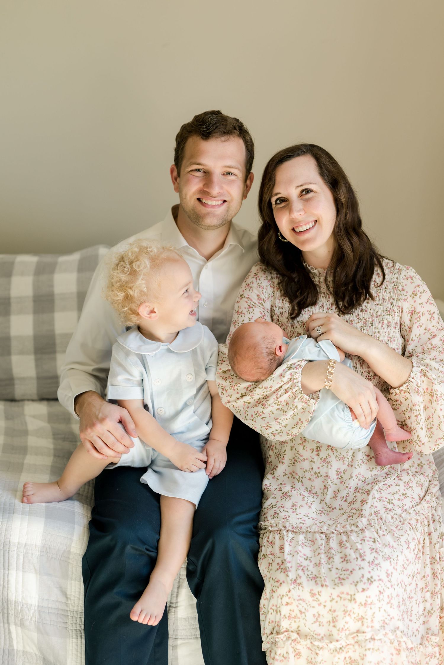Family of four sitting on couch together with toddler and newborn baby dressed in light blue outfits.