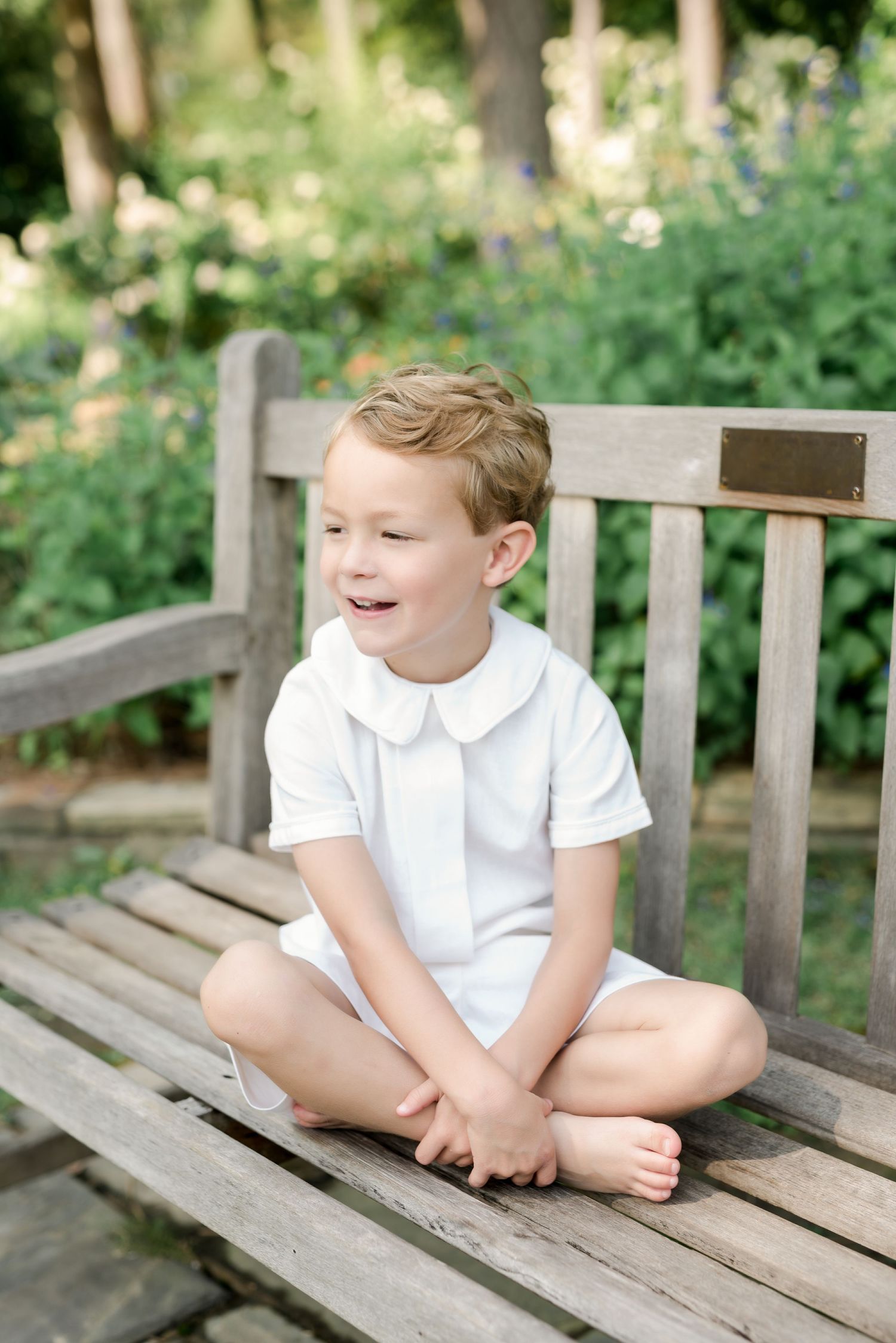 Child in white collar shirt sits casually on wooden bench in garden setting.