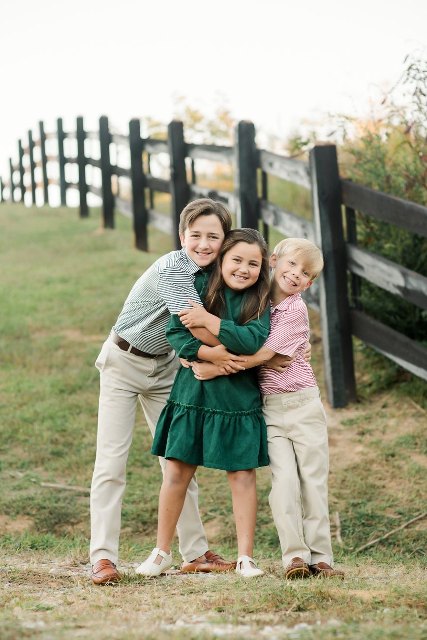 Three children hugging and smiling outdoors near a wooden fence on a grassy path during a family photo session.