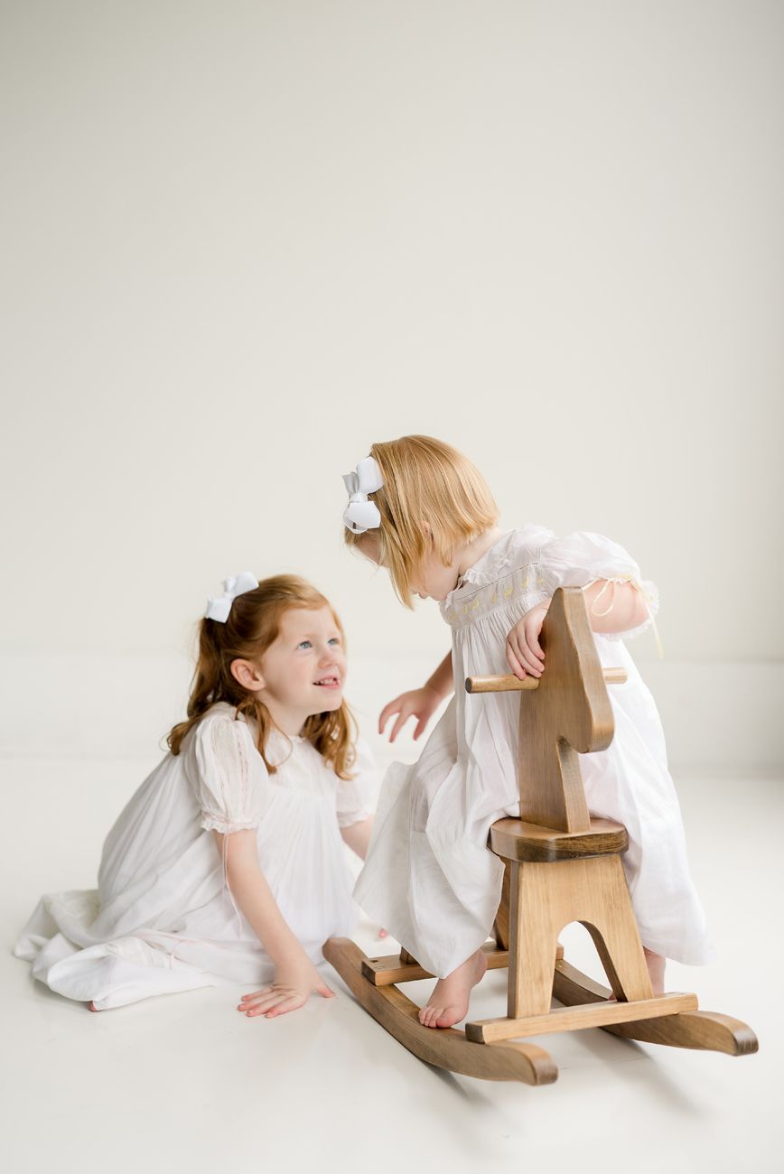 Two children in white dresses playing with wooden rocking toy against neutral backdrop.