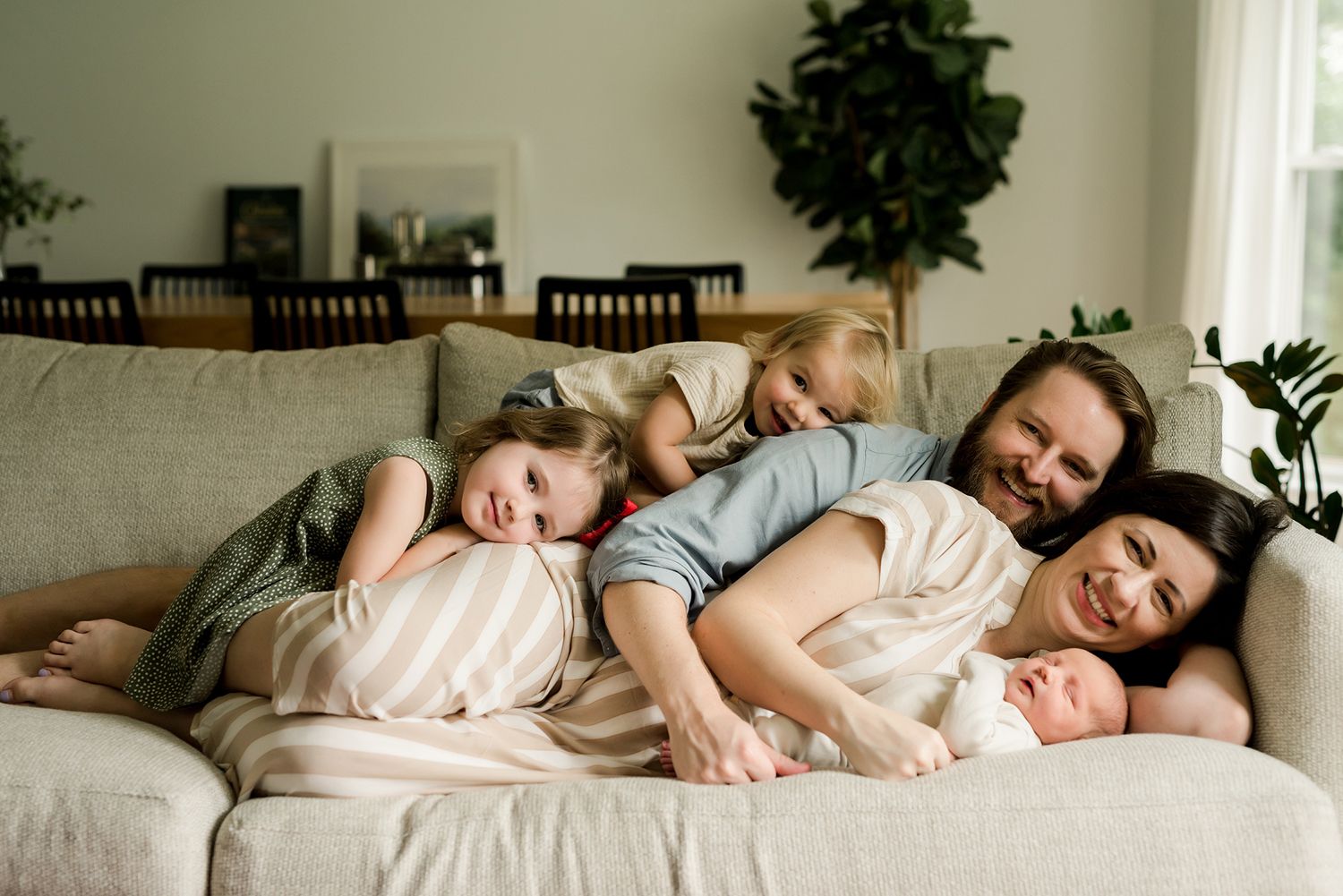 Family resting together on a beige couch with indoor plants and frames in the background.