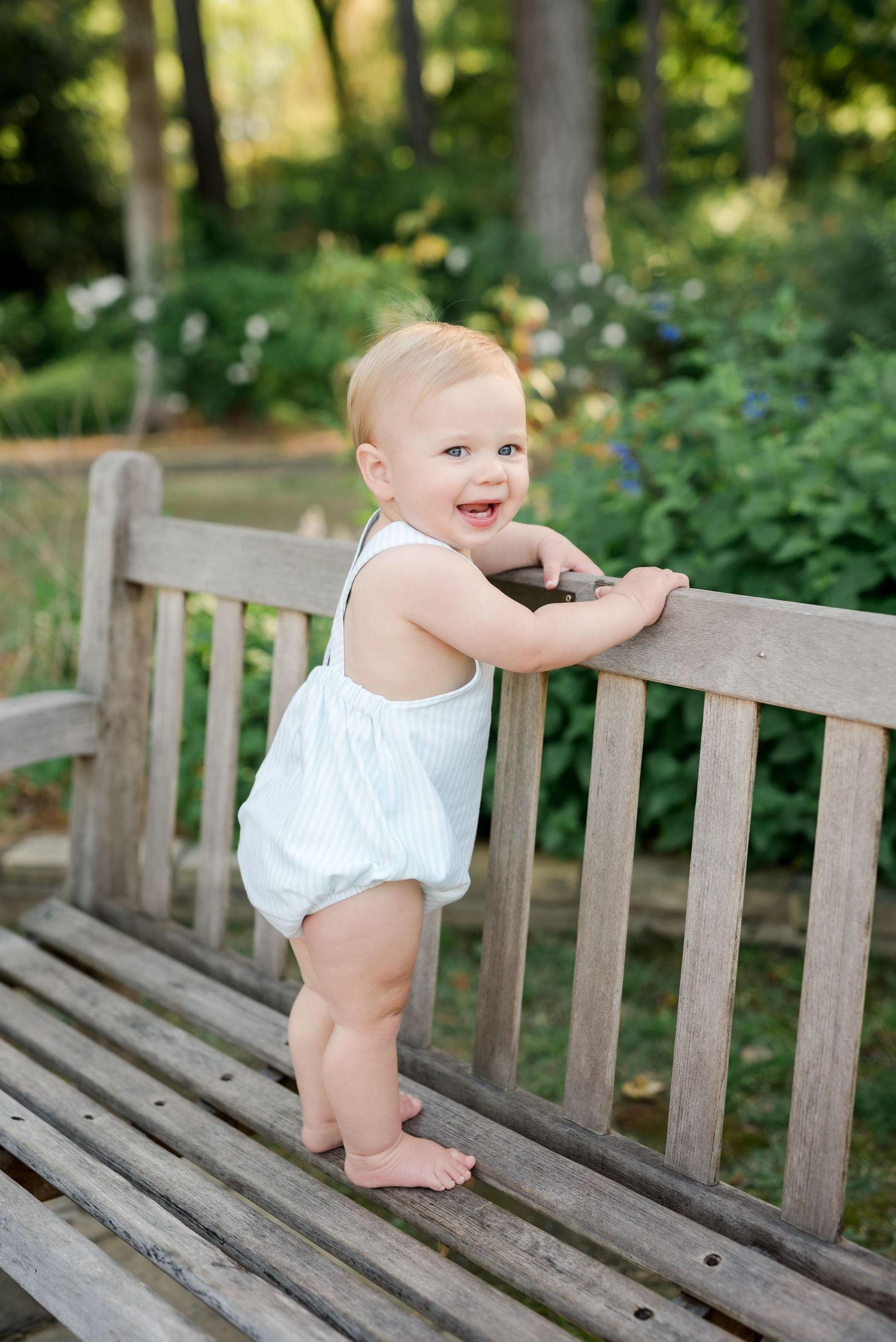 Baby in white romper standing and smiling at wooden garden bench.