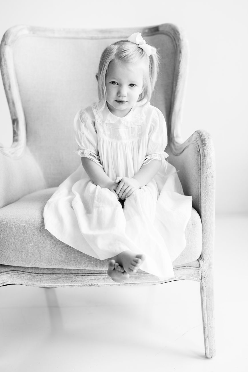 Child in vintage white dress sitting barefoot on gray armchair in black and white photo.