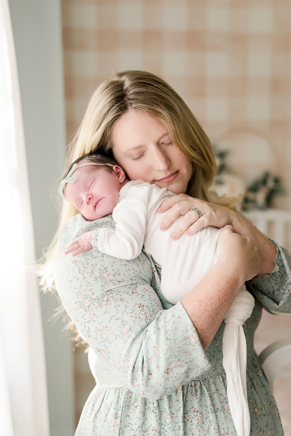 A gentle embrace between parent and newborn in a soft, light-filled room with floral wallpaper.