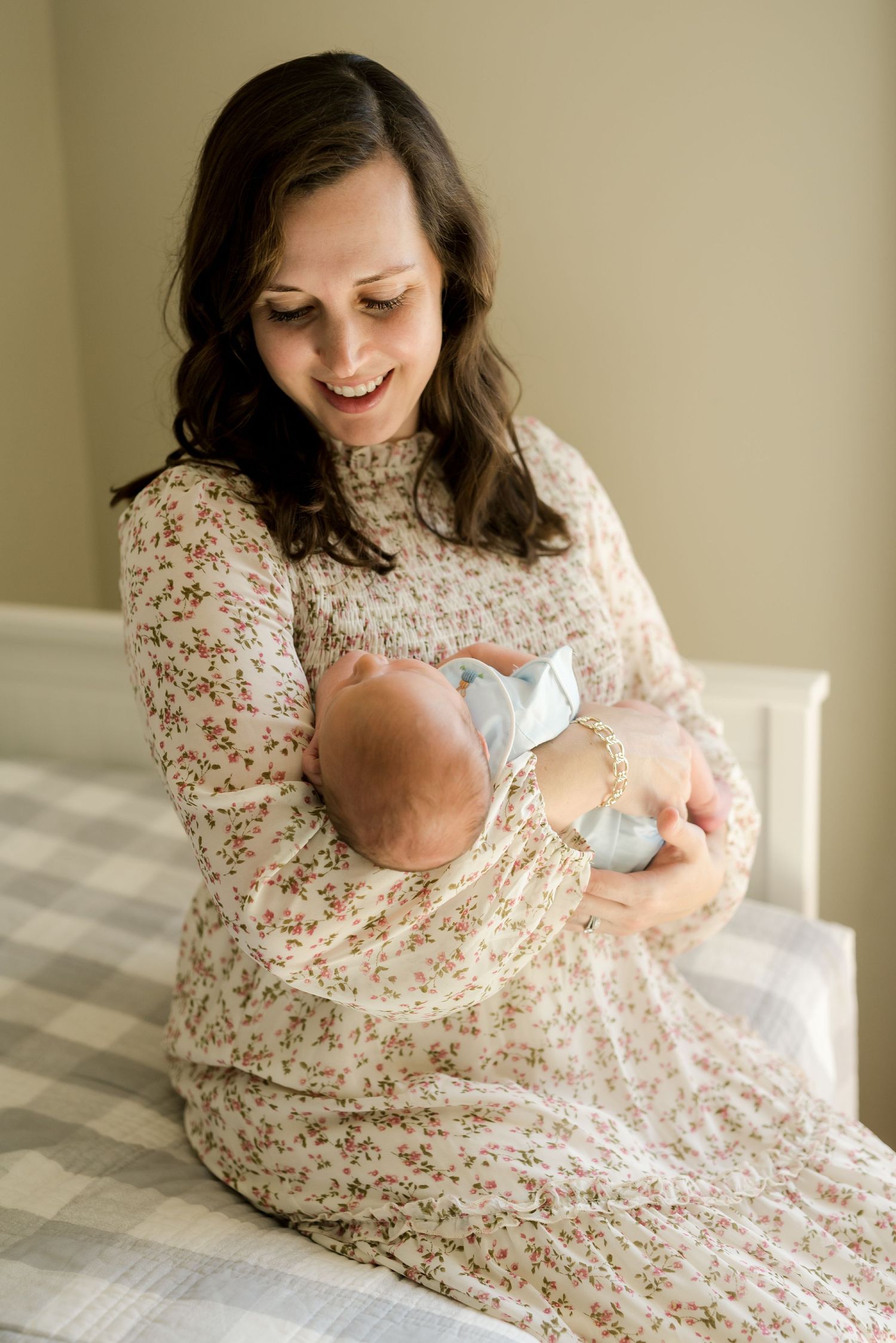 Woman in floral dress sitting on bed holding a sleeping baby.