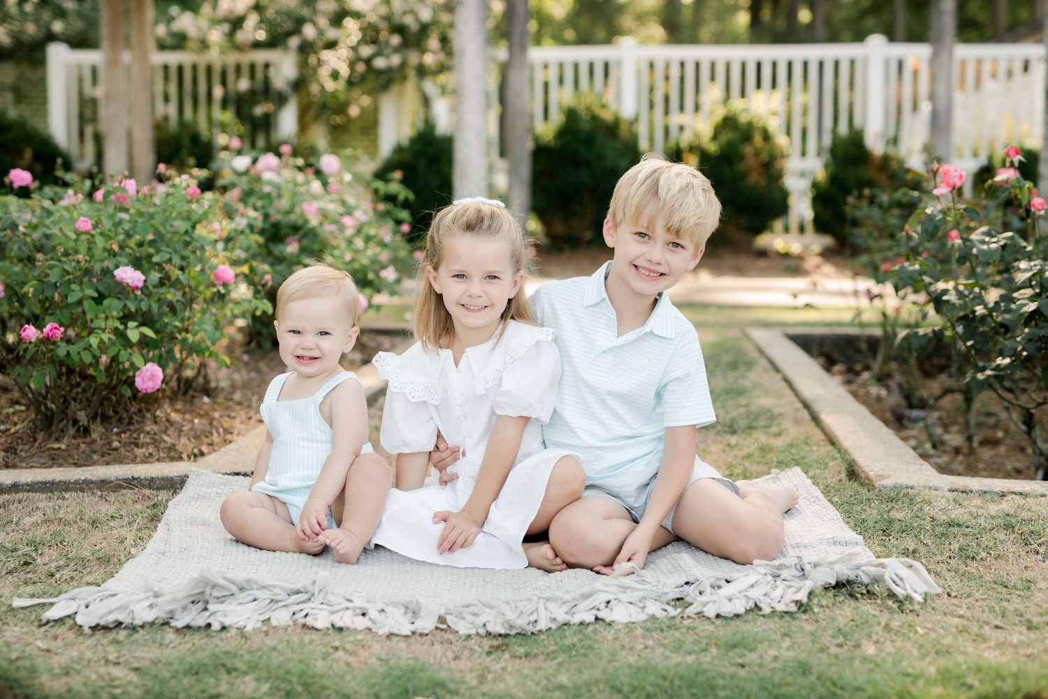 Three children in white summer outfits sitting together on blanket in garden setting.