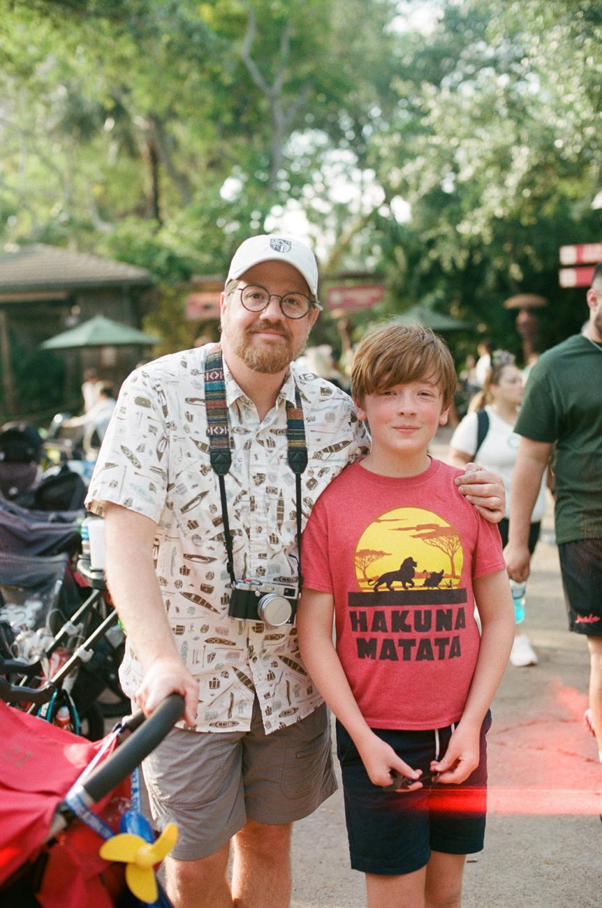 Two visitors in casual summer attire enjoying a day at a theme park, with one wearing a Hakuna Matata t-shirt.