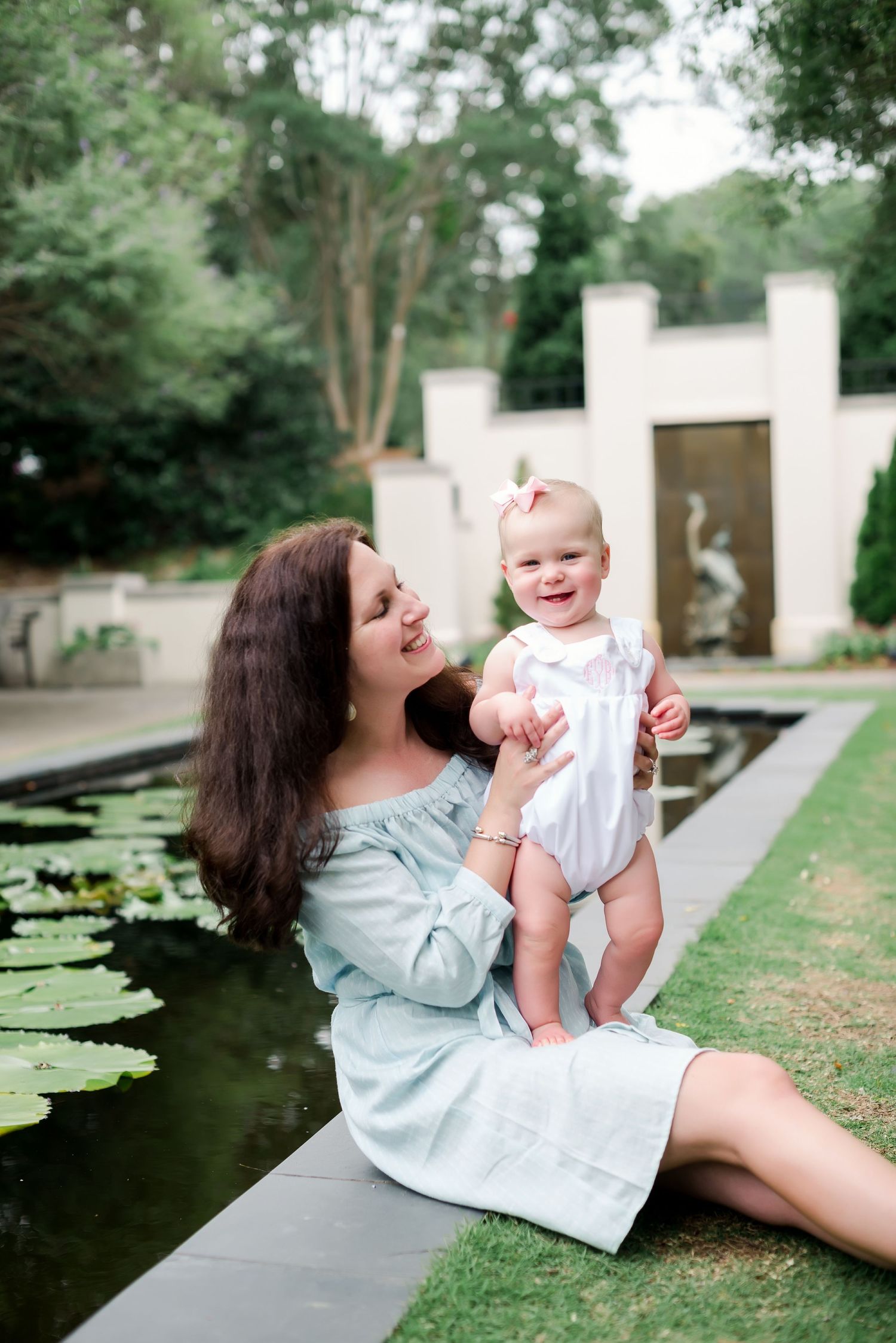 Sweet moment between mother and baby by a garden pond with lily pads and white architecture.