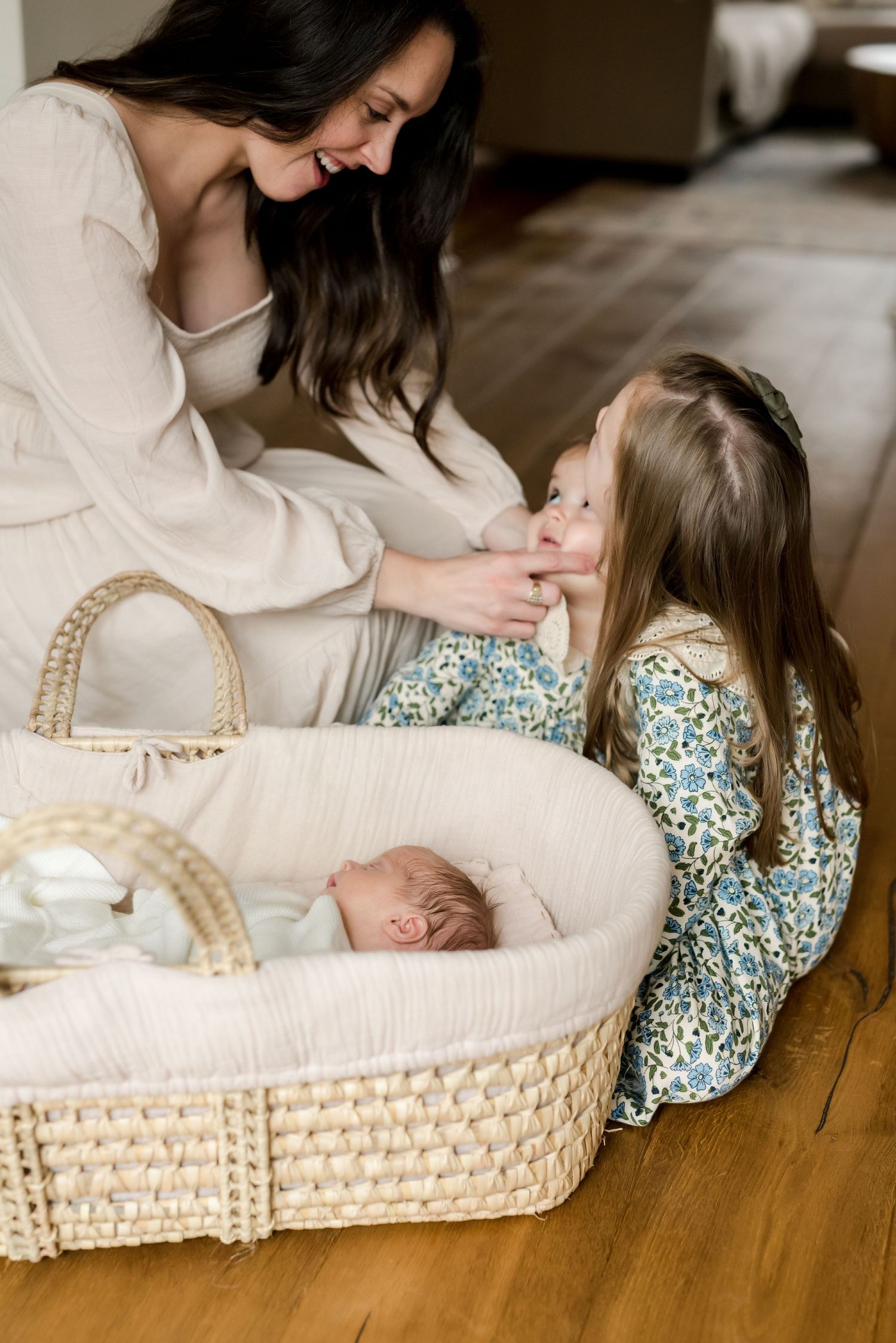 A tender moment between mother in cream sweater and child beside a woven bassinet with sleeping newborn.