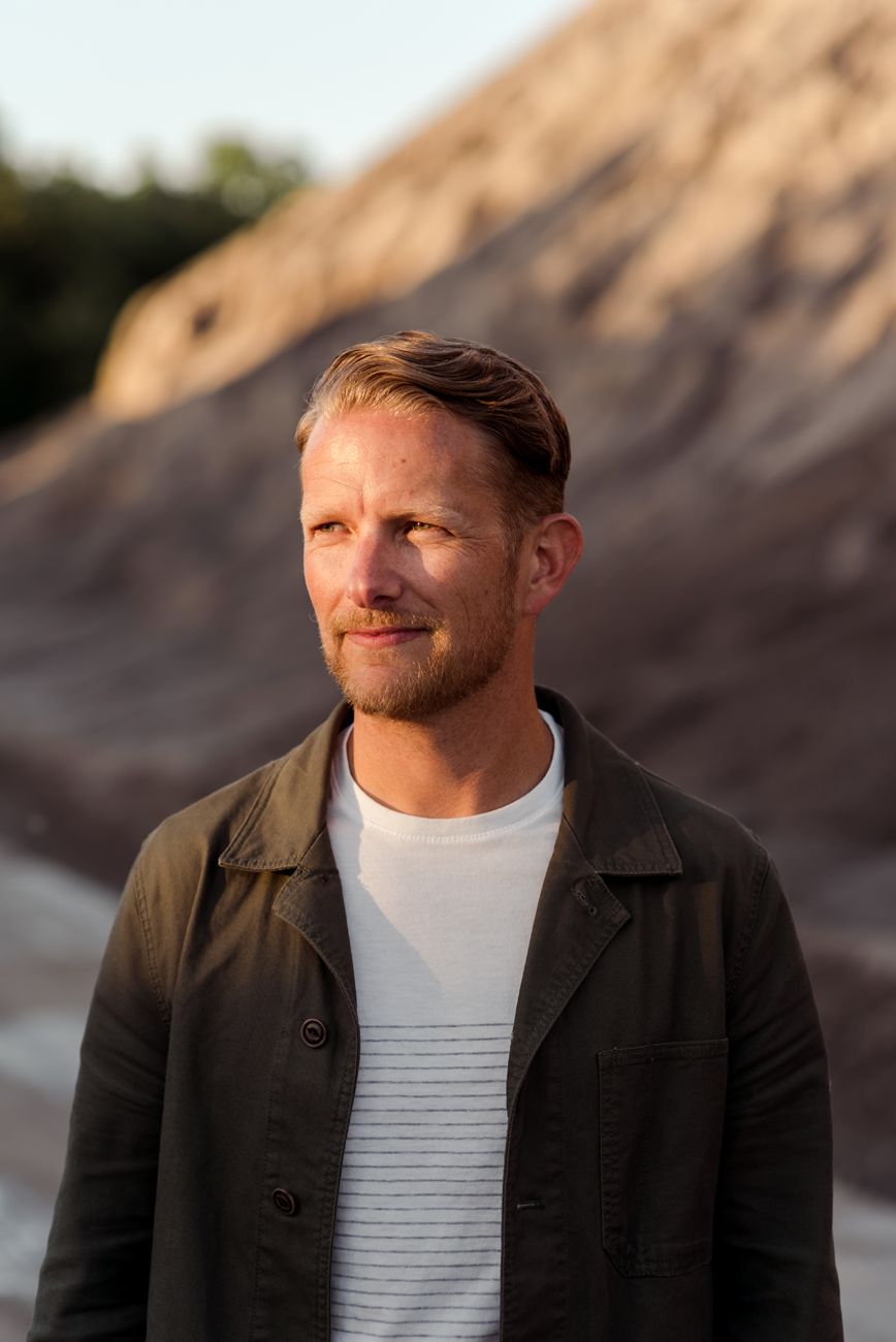 Person in dark jacket and white shirt poses against rocky landscape at sunset.