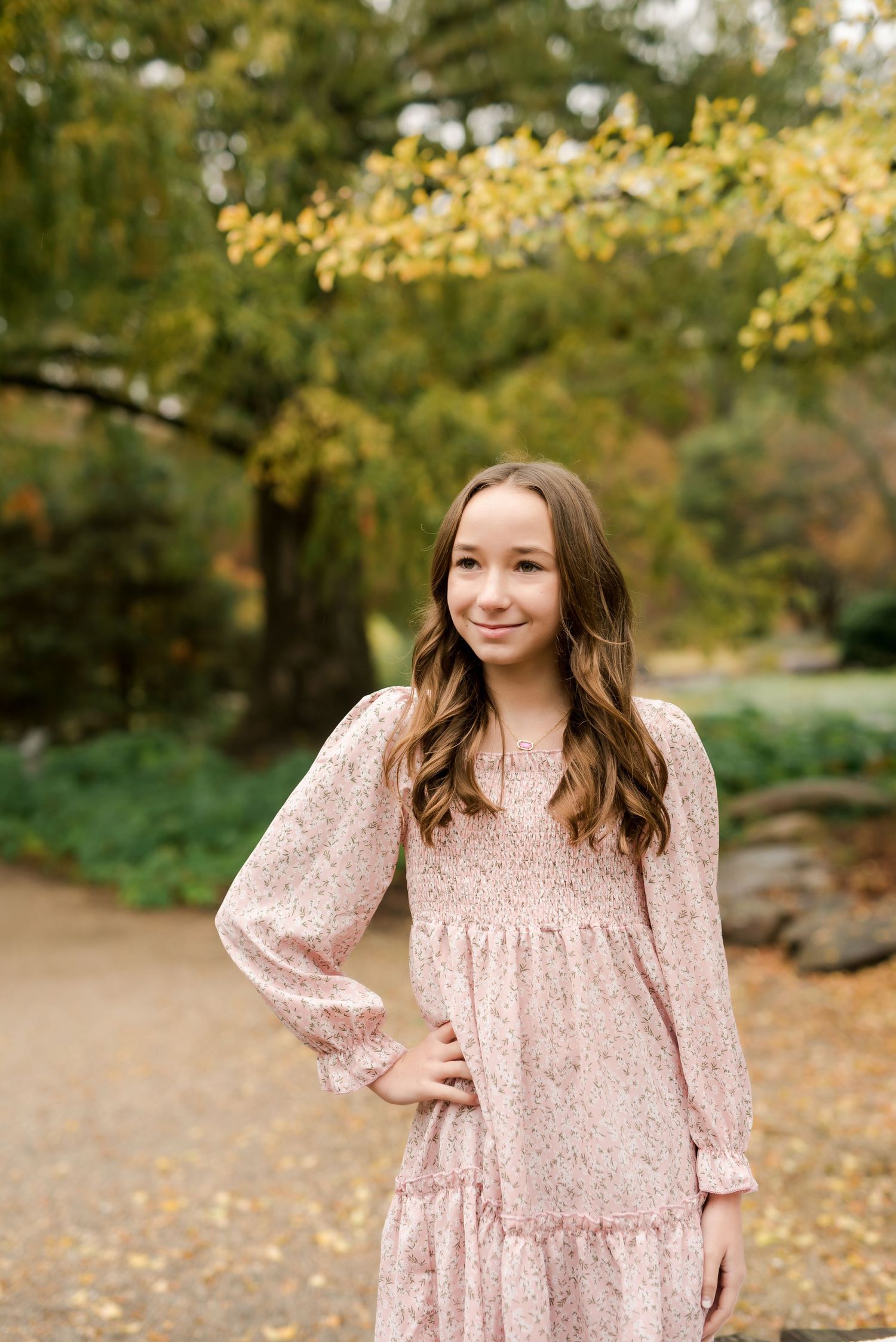 Young girl in pink floral dress poses outdoors among autumn foliage in a garden setting.