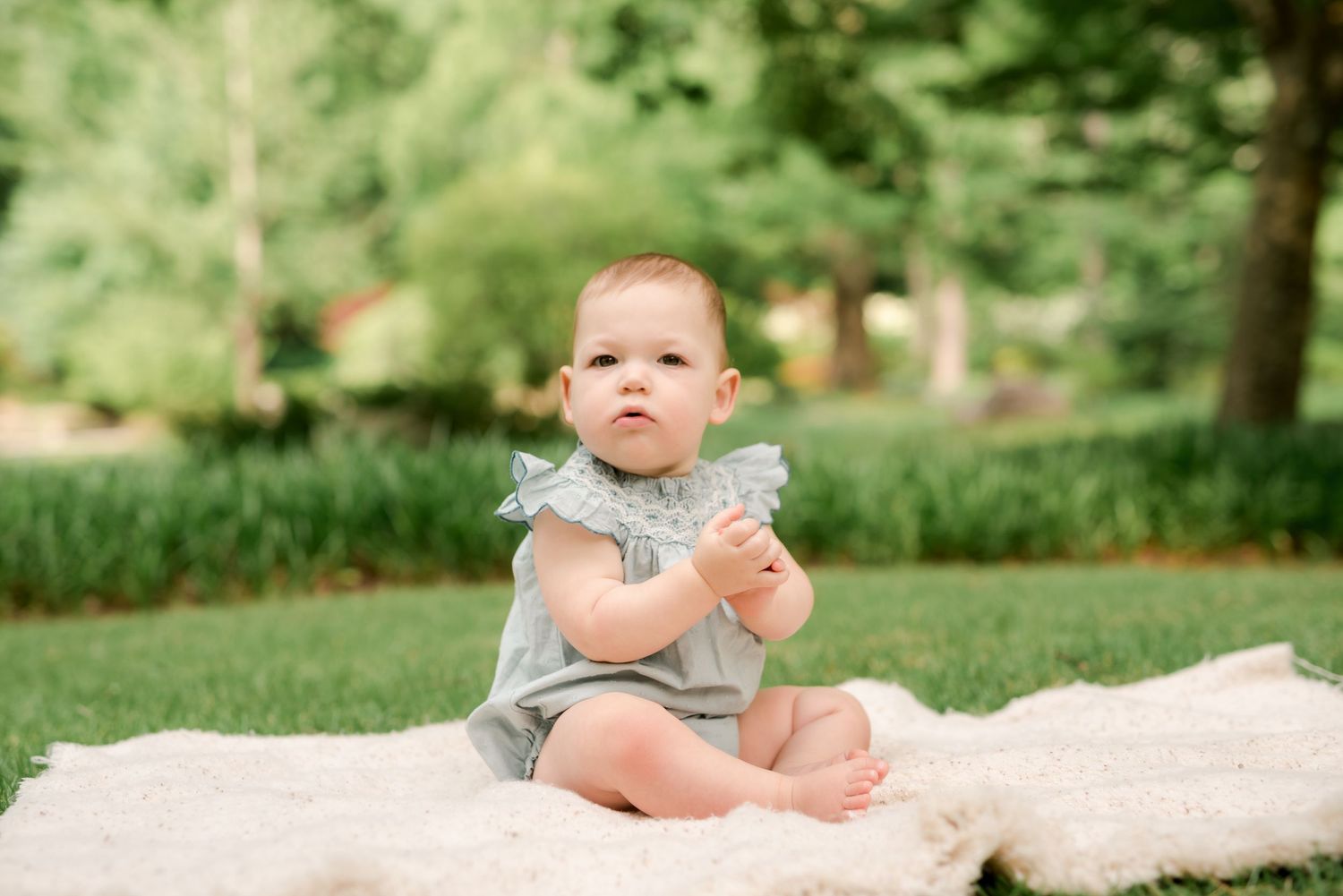 Baby in grey romper sitting on cream blanket in garden setting.