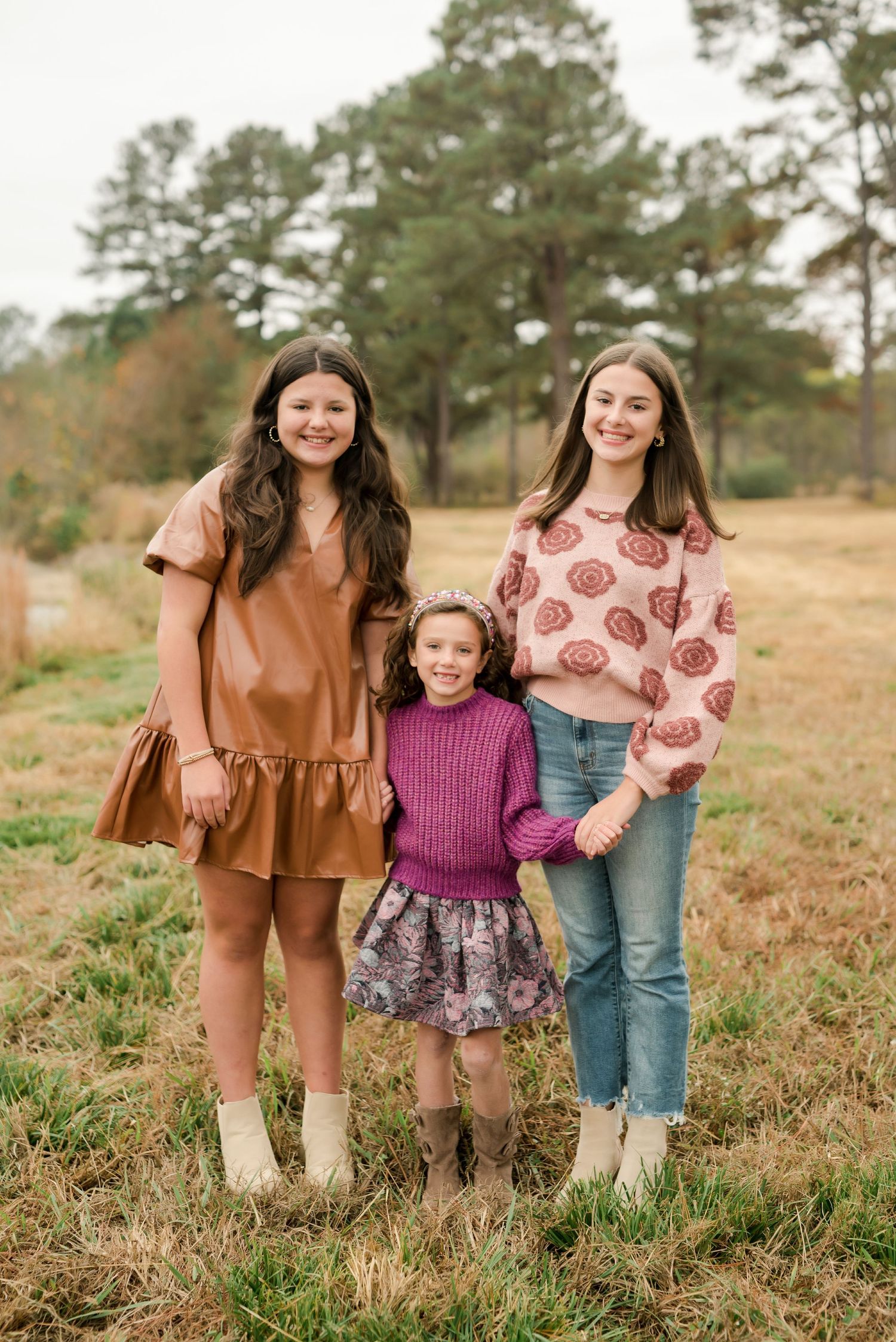 Three people in stylish fall outfits standing together in field with pine trees in background.