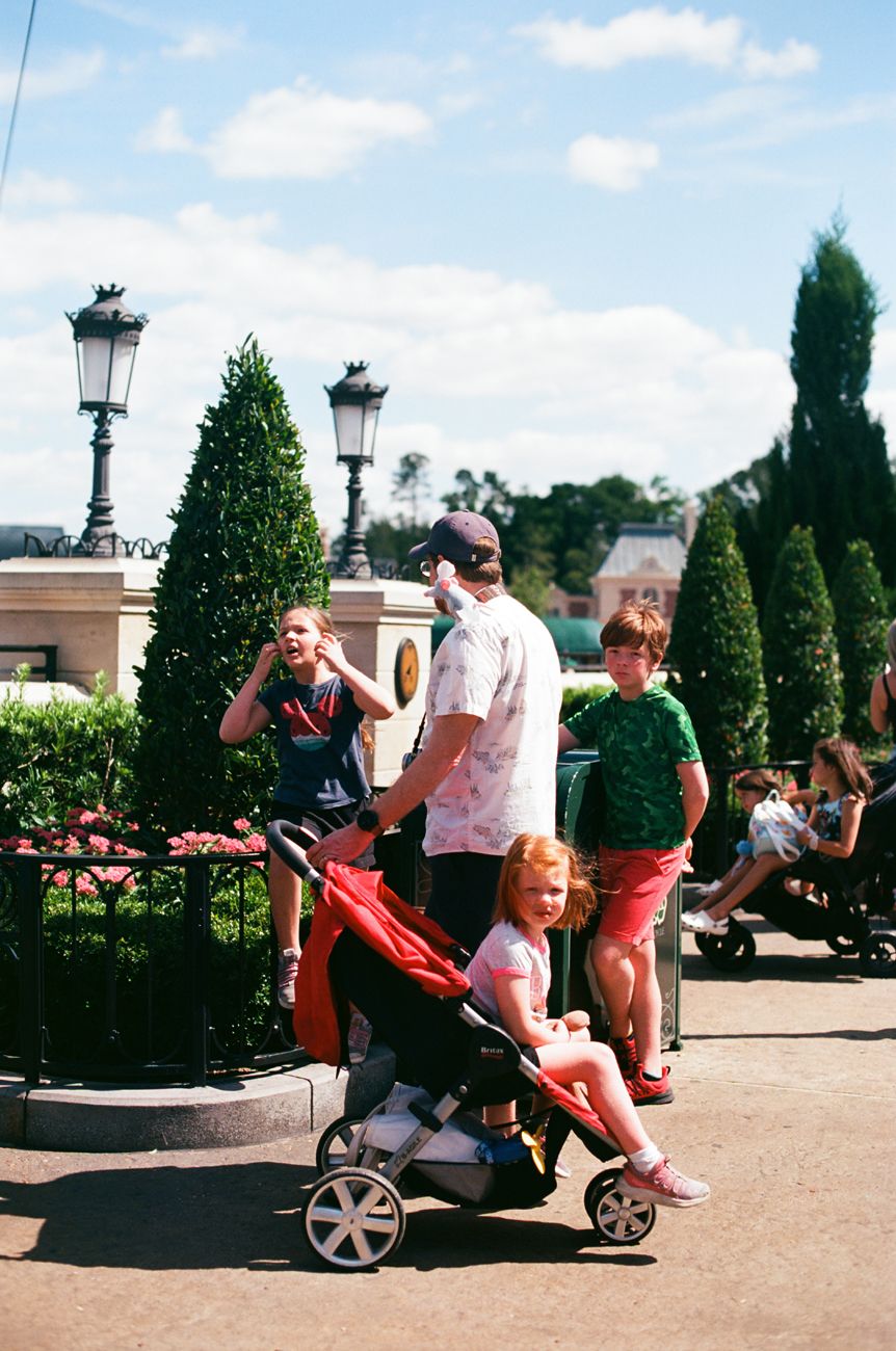 A family group stands near decorative lampposts and topiaries with a red stroller.