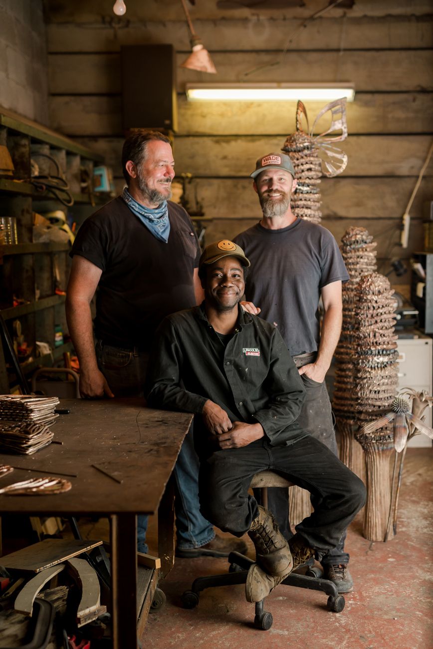 Three artisans pose together in workshop with decorative metal sculptures.