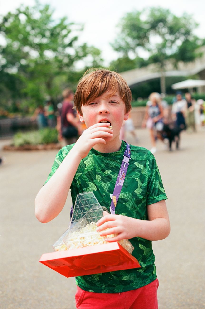 A young person in a green camouflage shirt enjoying popcorn from a red container at a theme park.