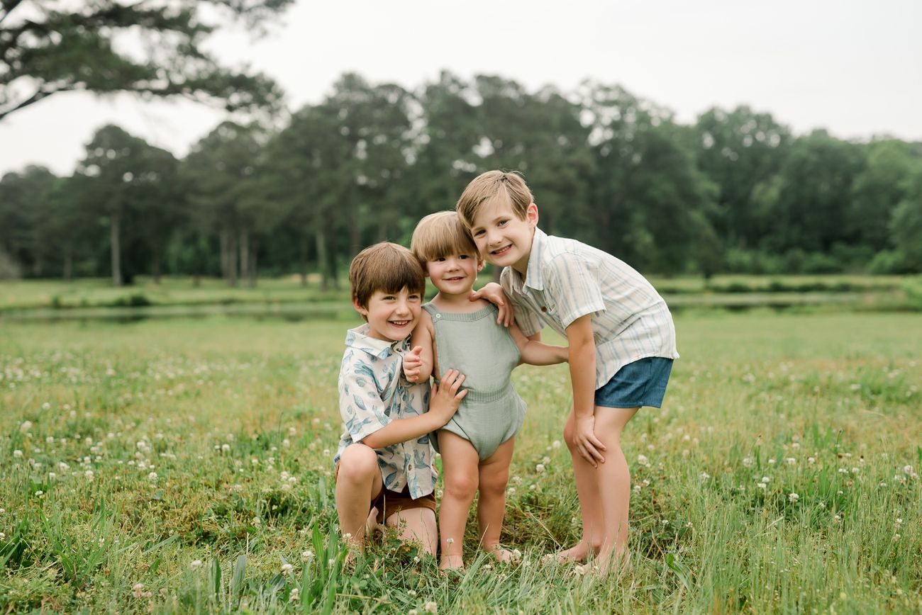 Three children playing and laughing together in a sunny field with pine trees in the background.