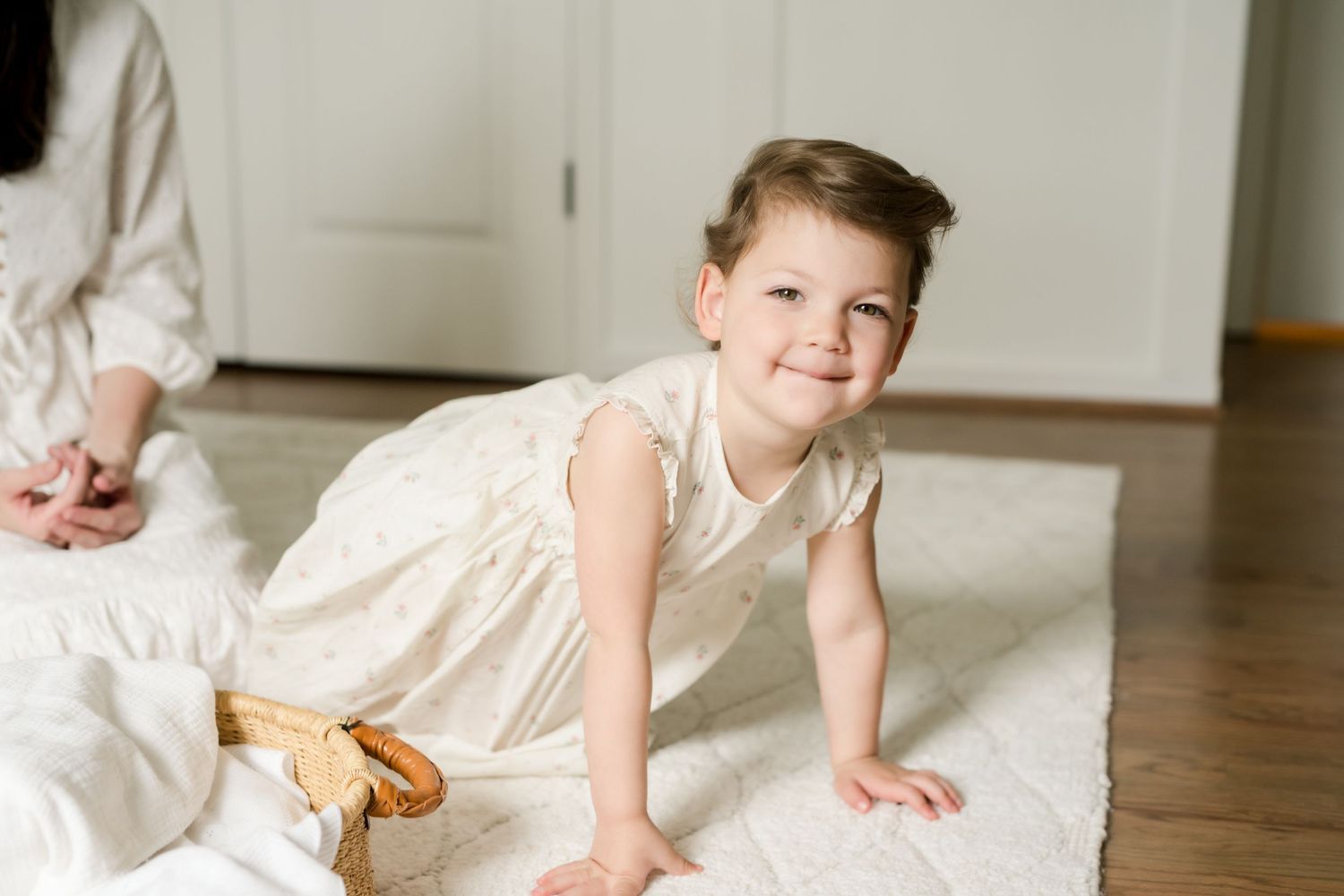 A child in cream-colored pajamas smiles while crawling on a white carpet on the floor.