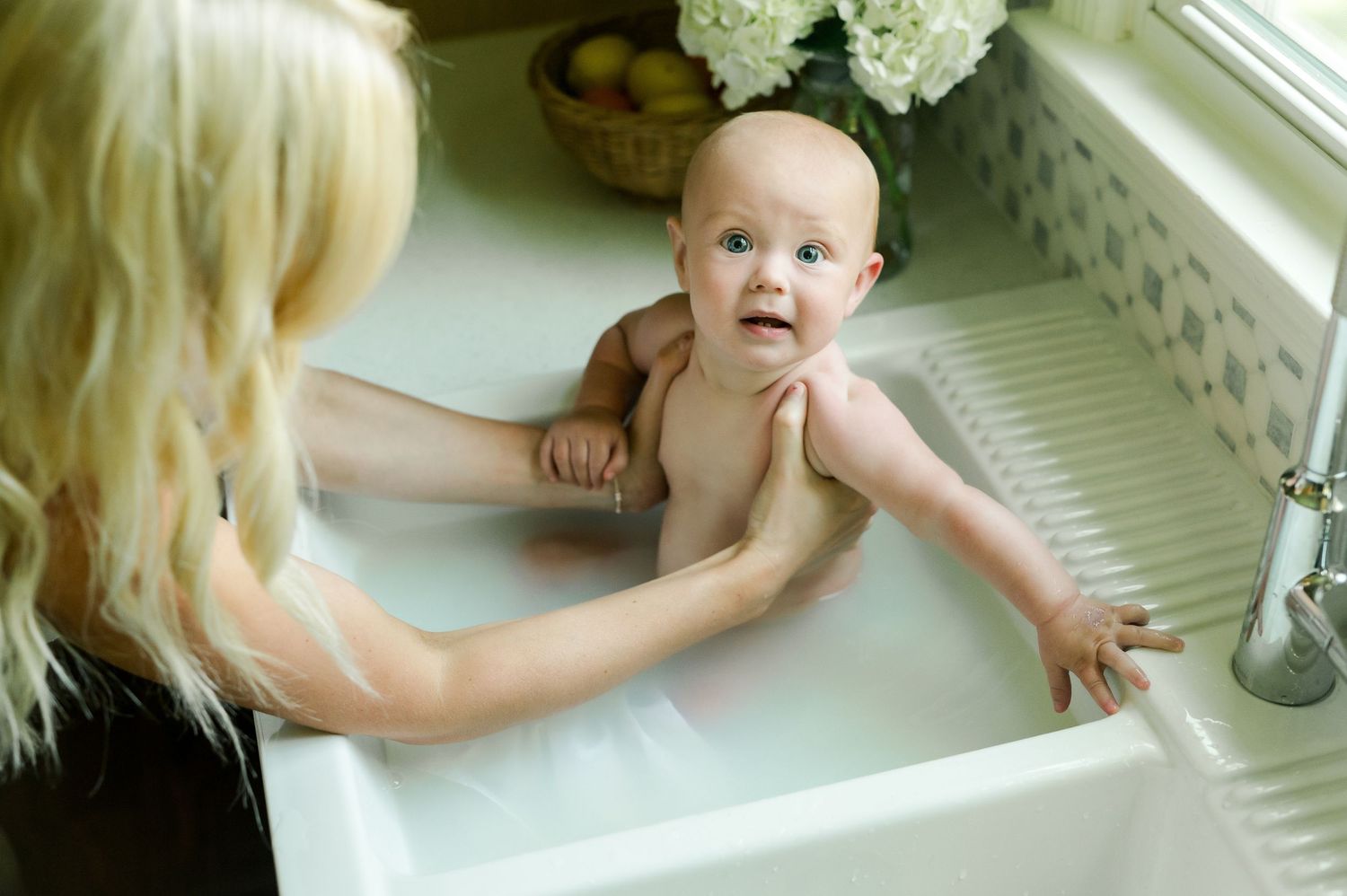 Parent bathing an infant in a white sink while supporting their head and body with gentle care.
