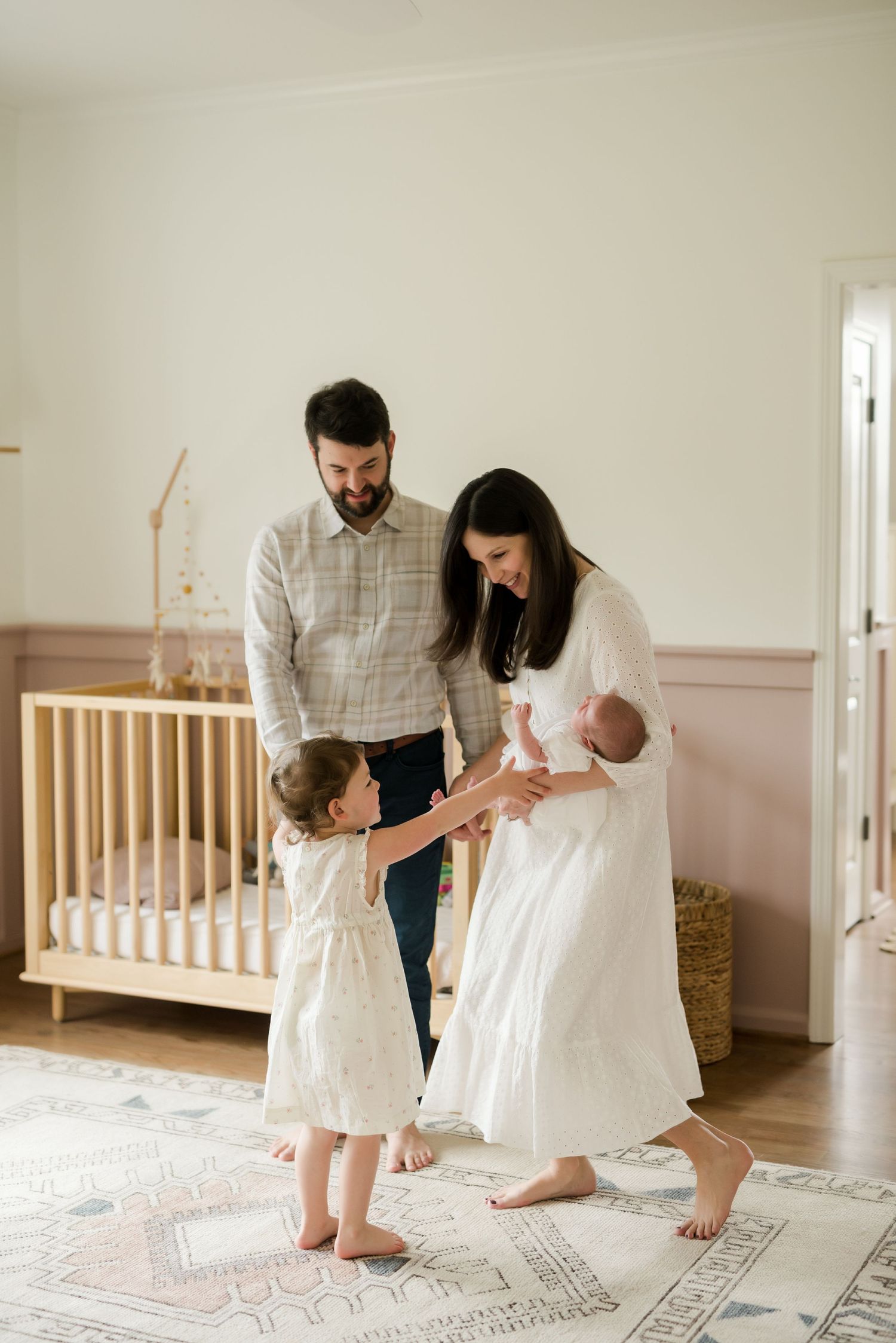 A family stands together in a nursery room with a wooden crib, soft area rug, and natural lighting.