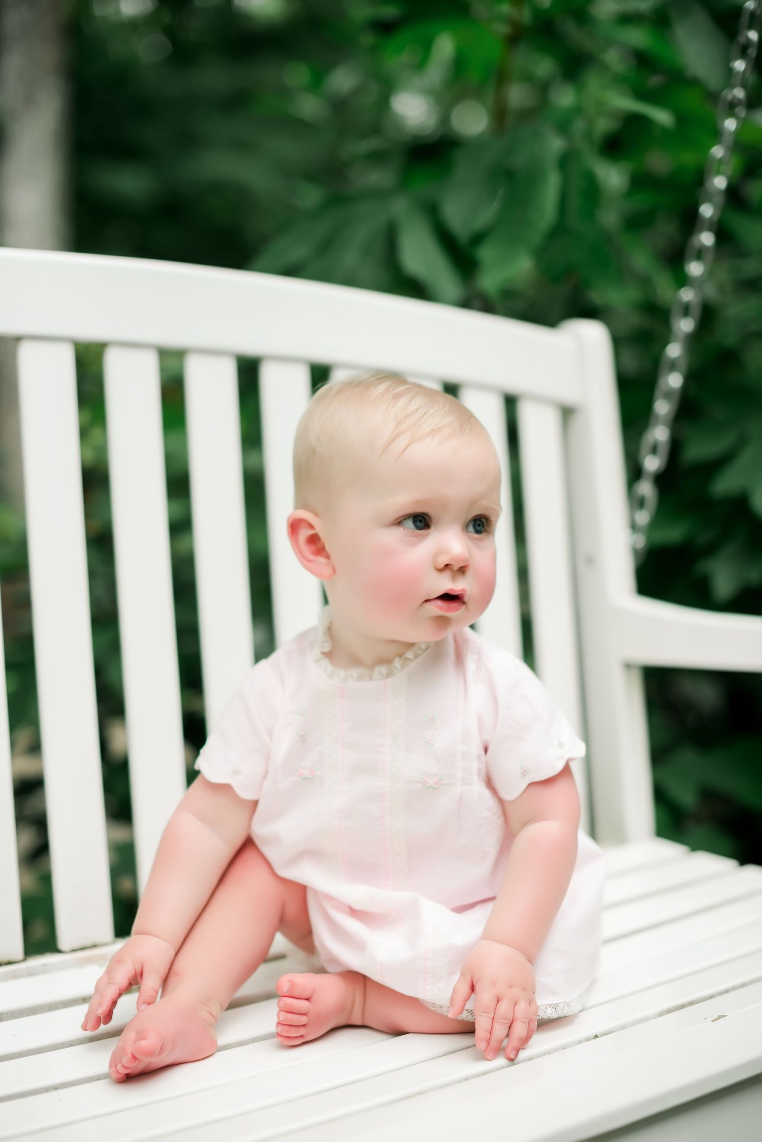Baby in white outfit sits on white wooden porch swing in garden setting.