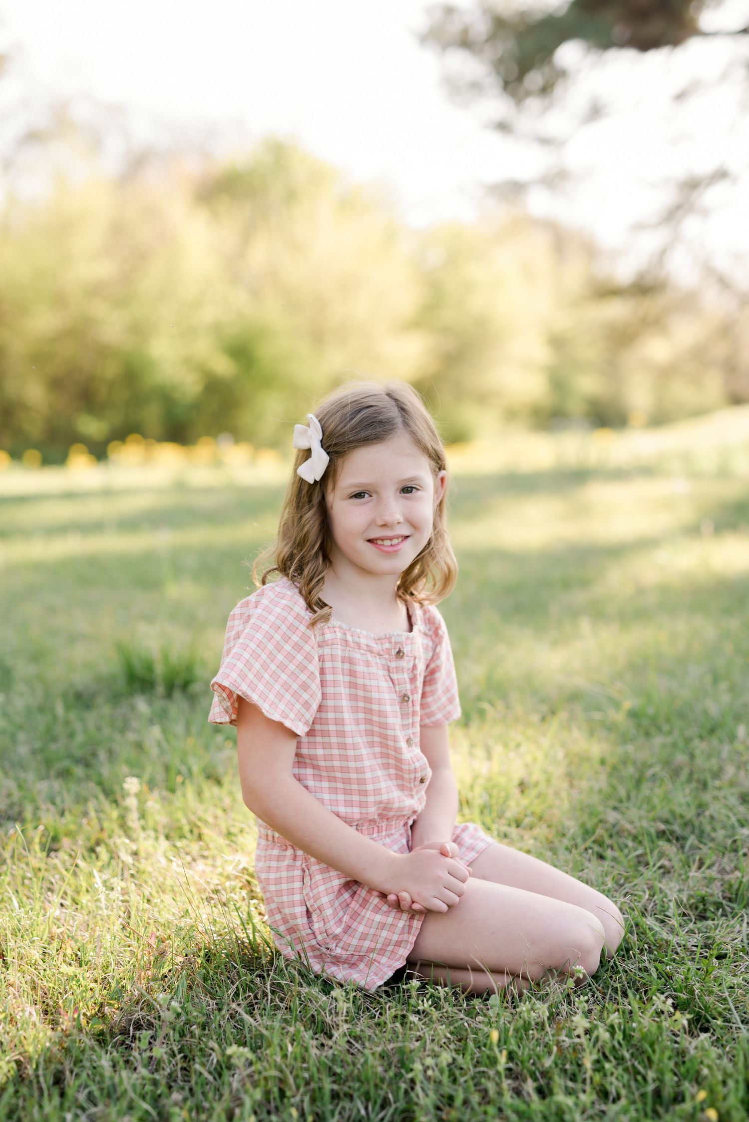 A smiling child in a pink gingham romper sits in a sunny meadow with a flower in their hair.